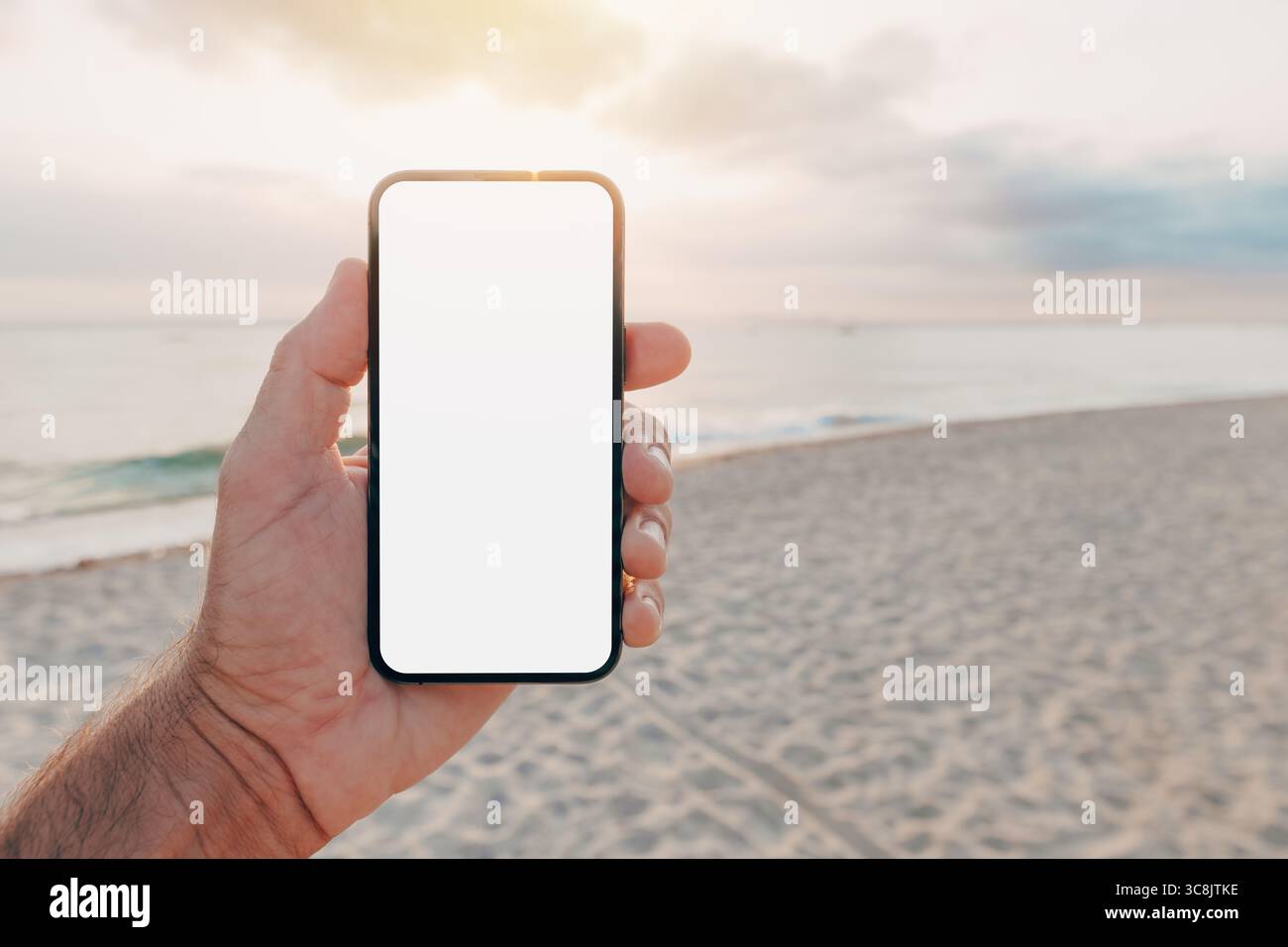 Handheld-Smartphone mit leerem Bildschirm am Strand bei Sonnenuntergang, App- oder Webinterface-Mockup bei natürlicher Außenbeleuchtung. Selektiver Fokus. Stockfoto