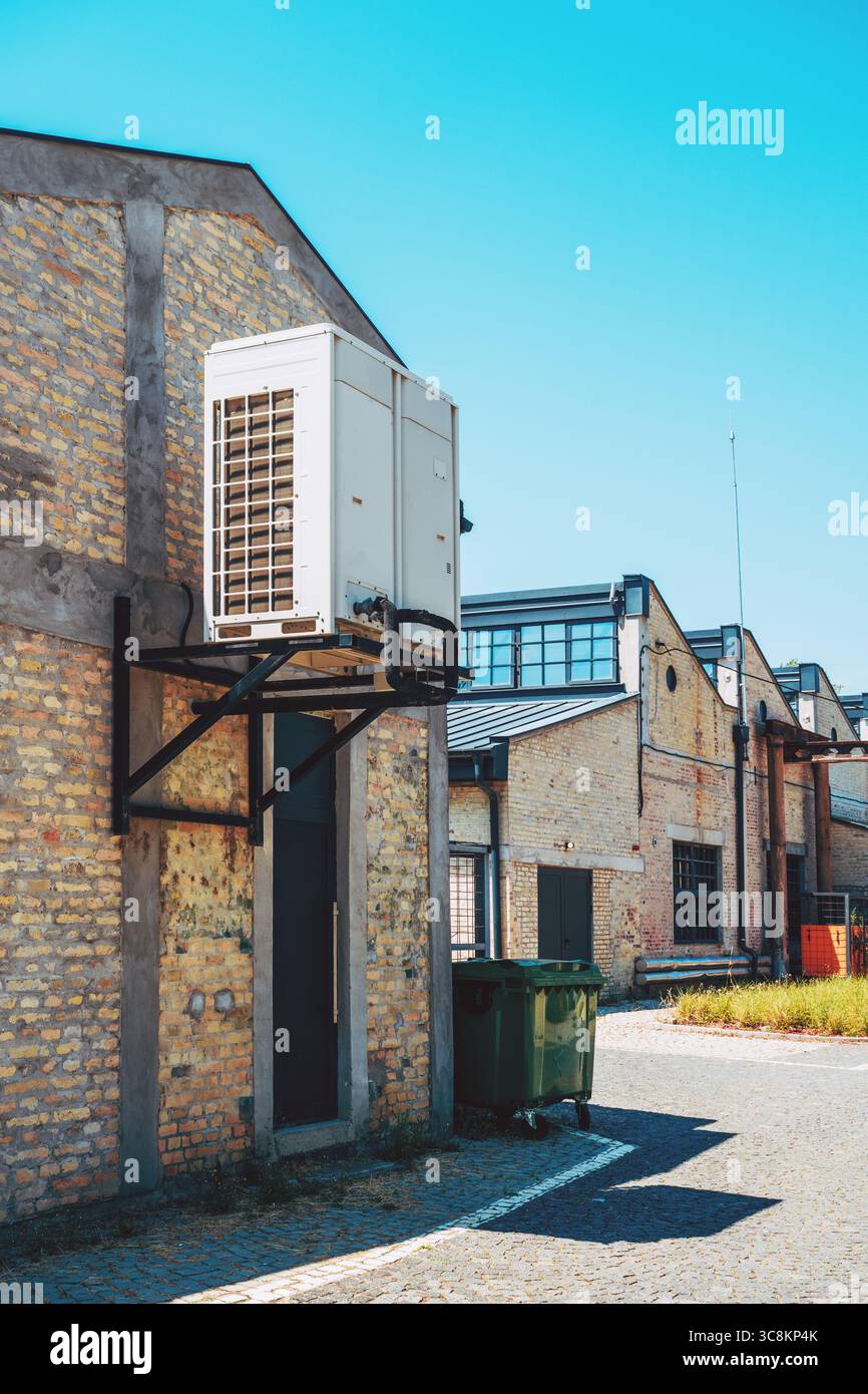 Wärmepumpeneinheit für den Außenbereich, montiert auf einem Industriegebäude aus Ziegelsteinen, mit blauem Himmel und sauberer Kopfsteinpflasterstraße, die HLK-Systeme in modernem Infrast repräsentiert Stockfoto