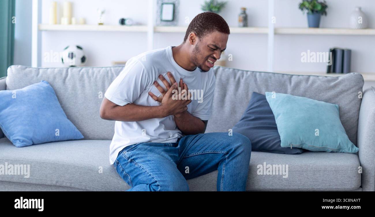 Millennial Afroamerikaner hat einen Herzinfarkt, berührt seine Brust vor Schmerzen, sitzt zu Hause auf dem Sofa Stockfoto