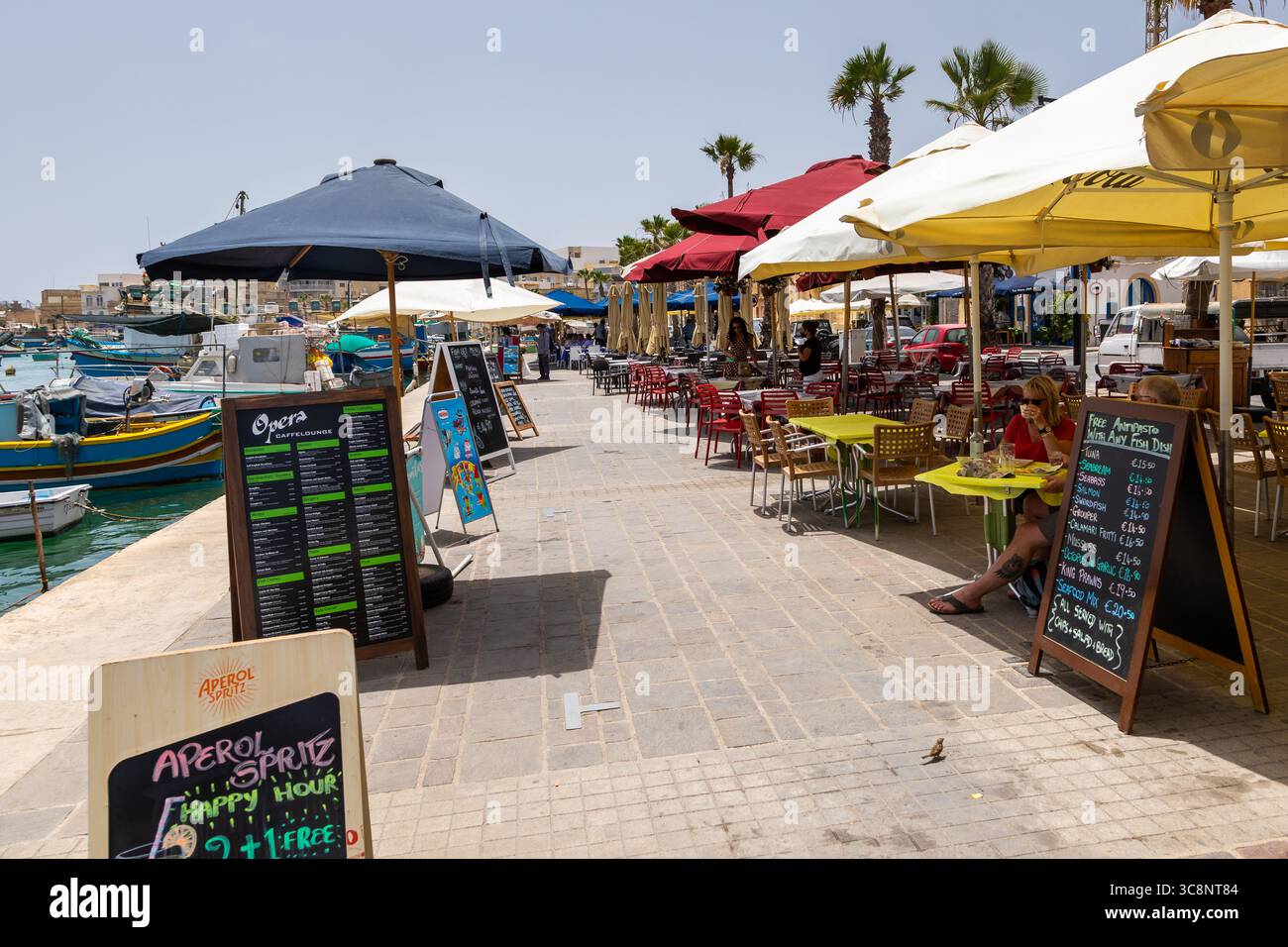 Marsaxlokk, Malta – 22. Juni 2021: Cafés und Restaurants mit Sitzgelegenheiten im Freien und farbenfrohen Sonnenschirmen entlang der Hafenpromenade. Stockfoto