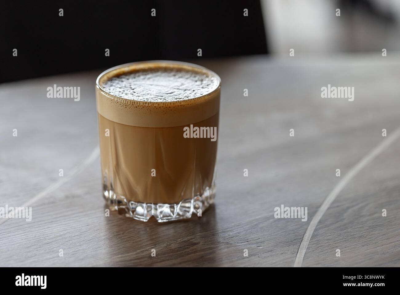 Flach weiß. Ein warmes, schaumiges Kaffeegetränk, serviert in einem klaren Glas auf einer rustikalen Tischplatte. Eine gemütliche und einladende Atmosphäre für die morgendliche Routine, entspannen Sie sich Stockfoto
