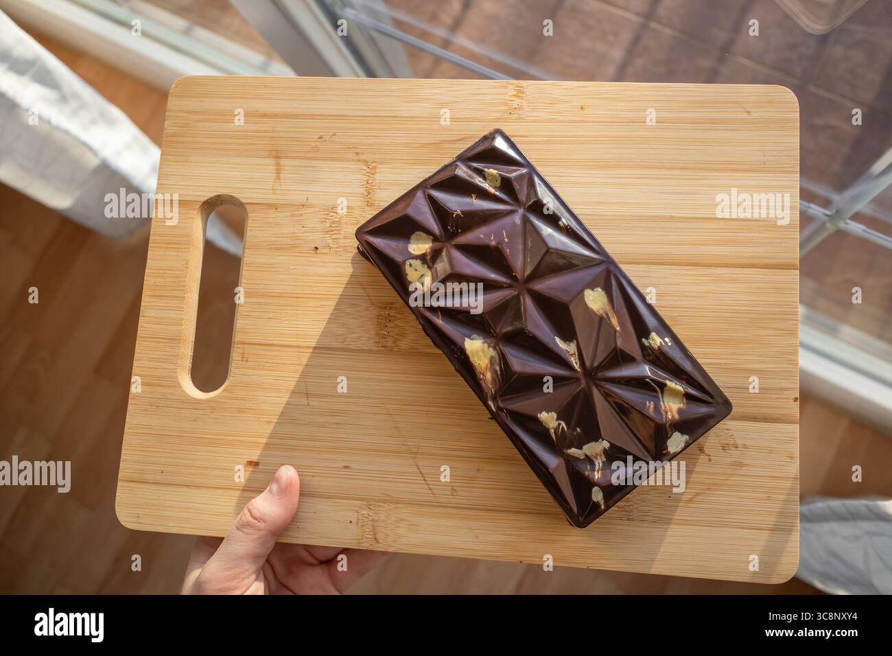 Menschliche Hand hält Dubai Schokolade auf hölzernem Schneidebrett vor dem Fenster. Wunderschöne Sweet Cocoa Bar bei Tageslicht. Stockfoto