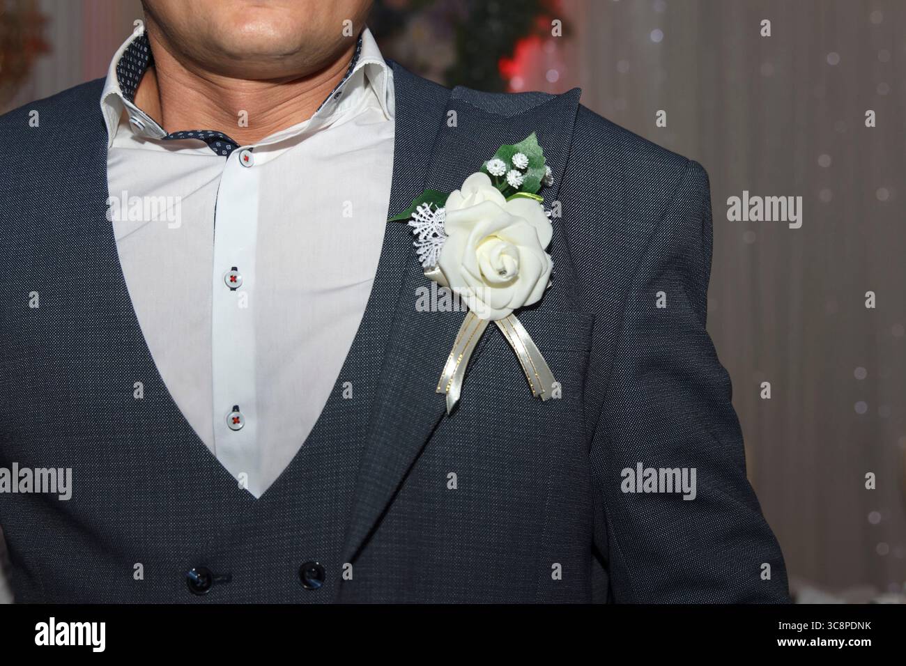 Ein Mann in einem formellen Anzug mit einer weißen Blumenkuppe. Der Hintergrund ist leicht verschwommen, was auf eine festliche oder feierliche Atmosphäre hindeutet Stockfoto