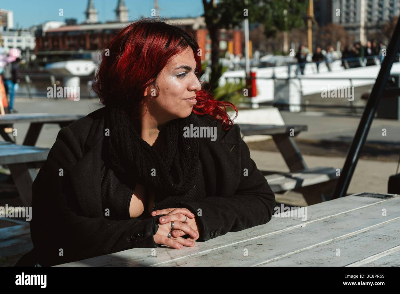 Junge Frau mit gefärbten roten Haaren und schwarzem Mantel und Schal, die an einem Tisch im Freien in buenos aires, argentinien, sitzt Stockfoto