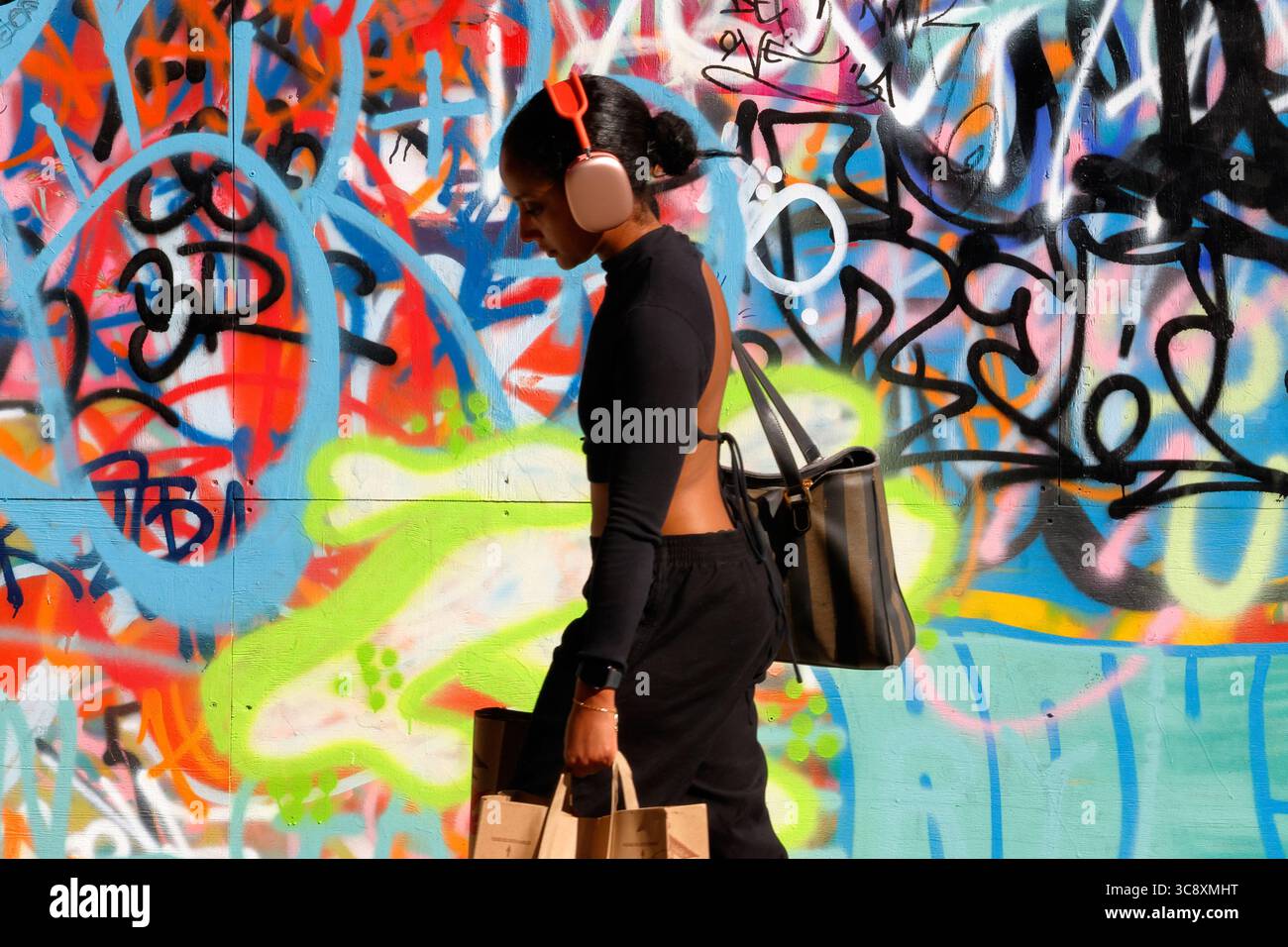 Eine junge Frau mit Kopfhörergesicht, teilweise im Schatten, mit Einkaufstaschen, die vor einem lebhaften Graffiti-Hintergrund auf den Boden schauen. Stockfoto