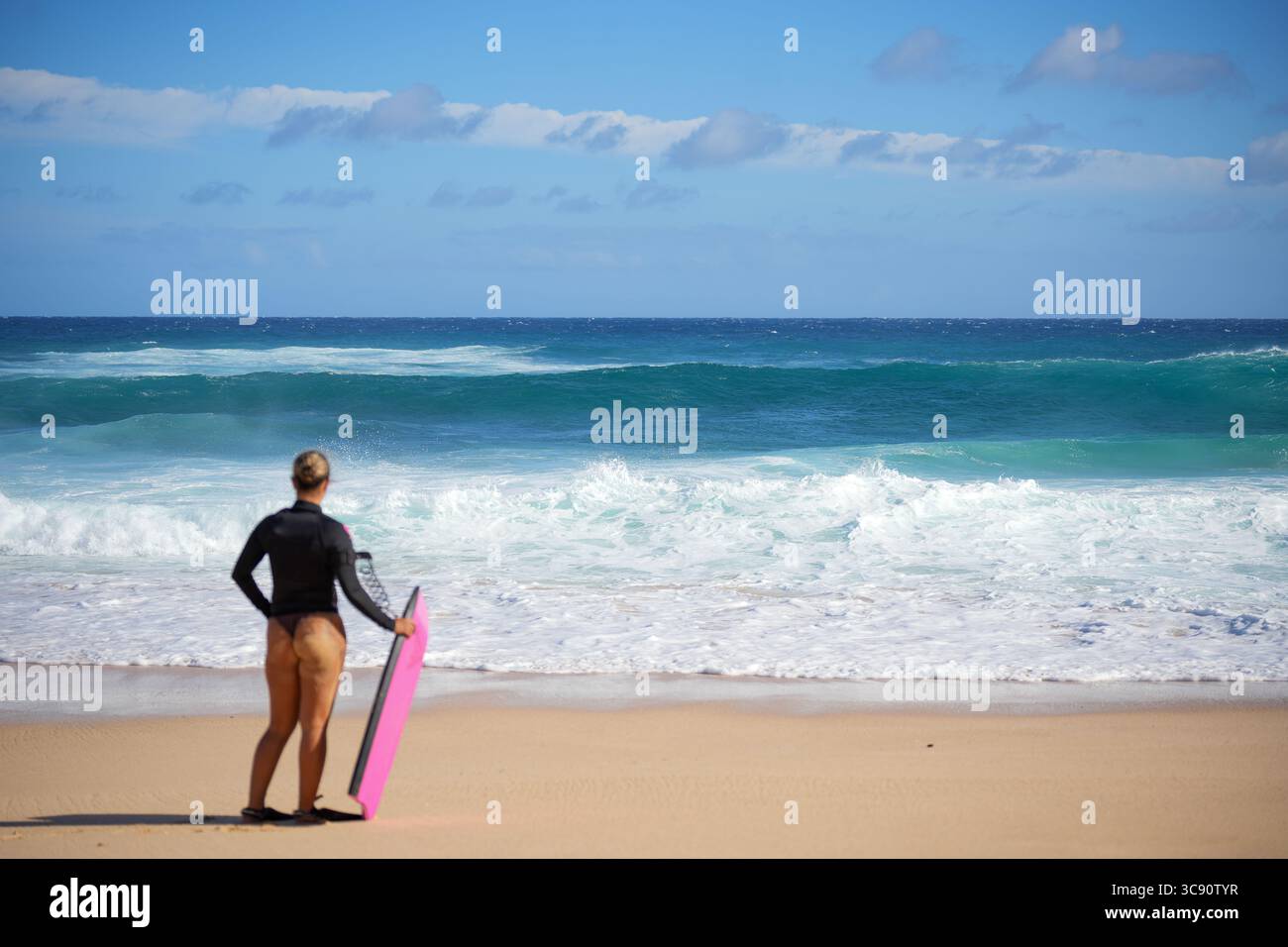 Bodyboarder steht am stürmischen Strand, hält das Bodyboard aufrecht im Sand und blickt auf die große Meeressturmwelle – zwischen Angst und Nervenkitzel, mit Blick auf das Meer Stockfoto