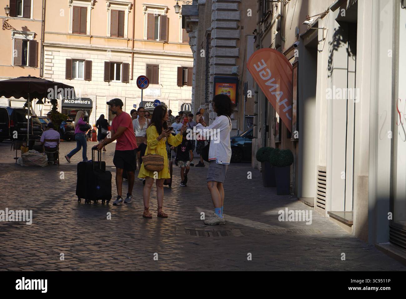 Rom, Italien, 13. Juli 2025: Ein Mann fotografiert eine Frau in gelbem Kleid mit Eis auf einer Kopfsteinpflasterstraße und hält einen klassischen römischen Moment fest. Stockfoto