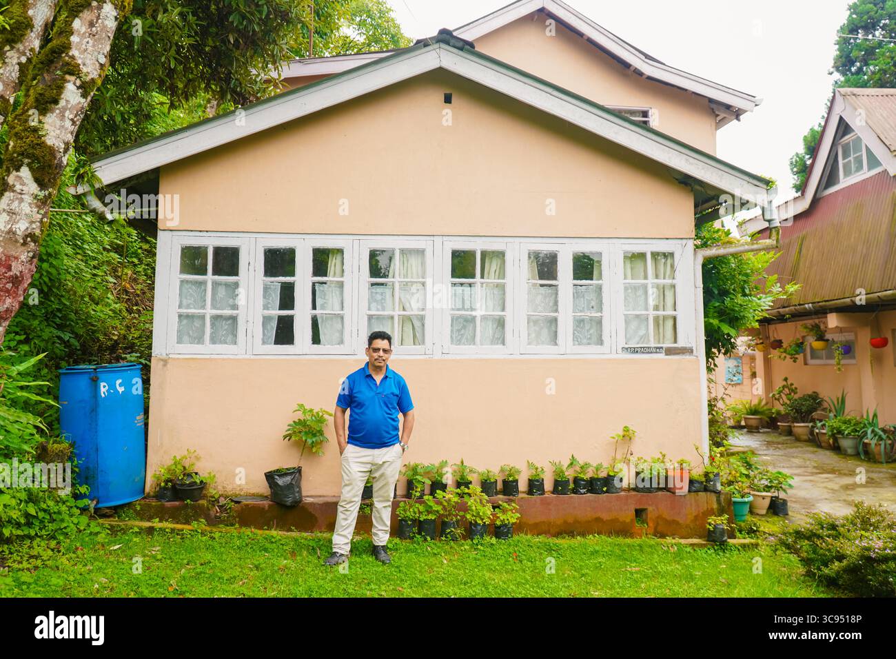 Mann steht vor einem traditionellen Landhaus mit weißen Fenstern und Garten in üppiger grüner Umgebung. Stockfoto