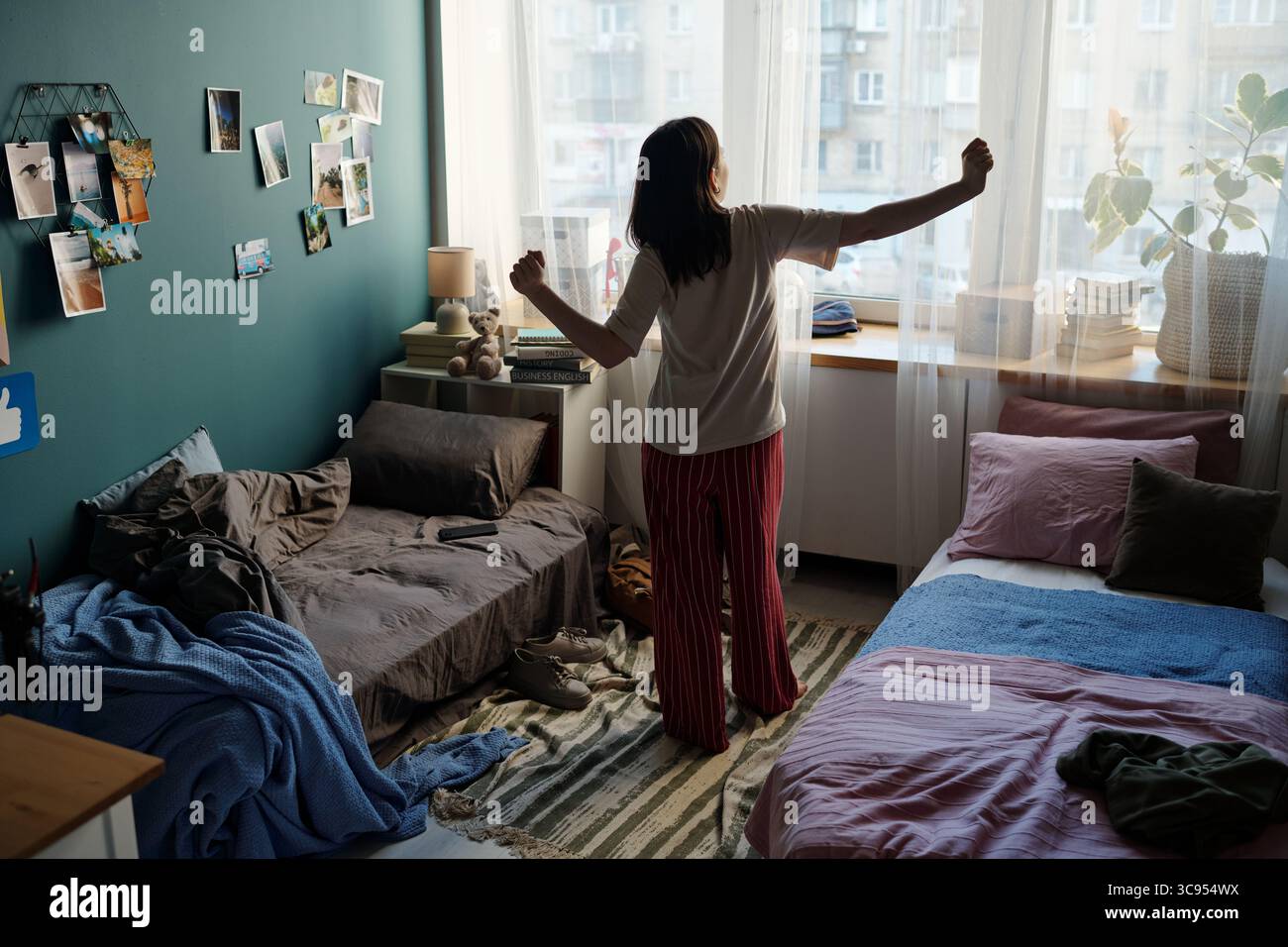 Junge Erwachsene kaukasische Frau, die neben dem Fenster steht und Arme in einem gemeinsamen Schlafzimmer mit zwei Betten ausdehnt, Morgenlicht durch transparente Vorhänge, persönliche Gegenstände auf dem Boden verstreut Stockfoto