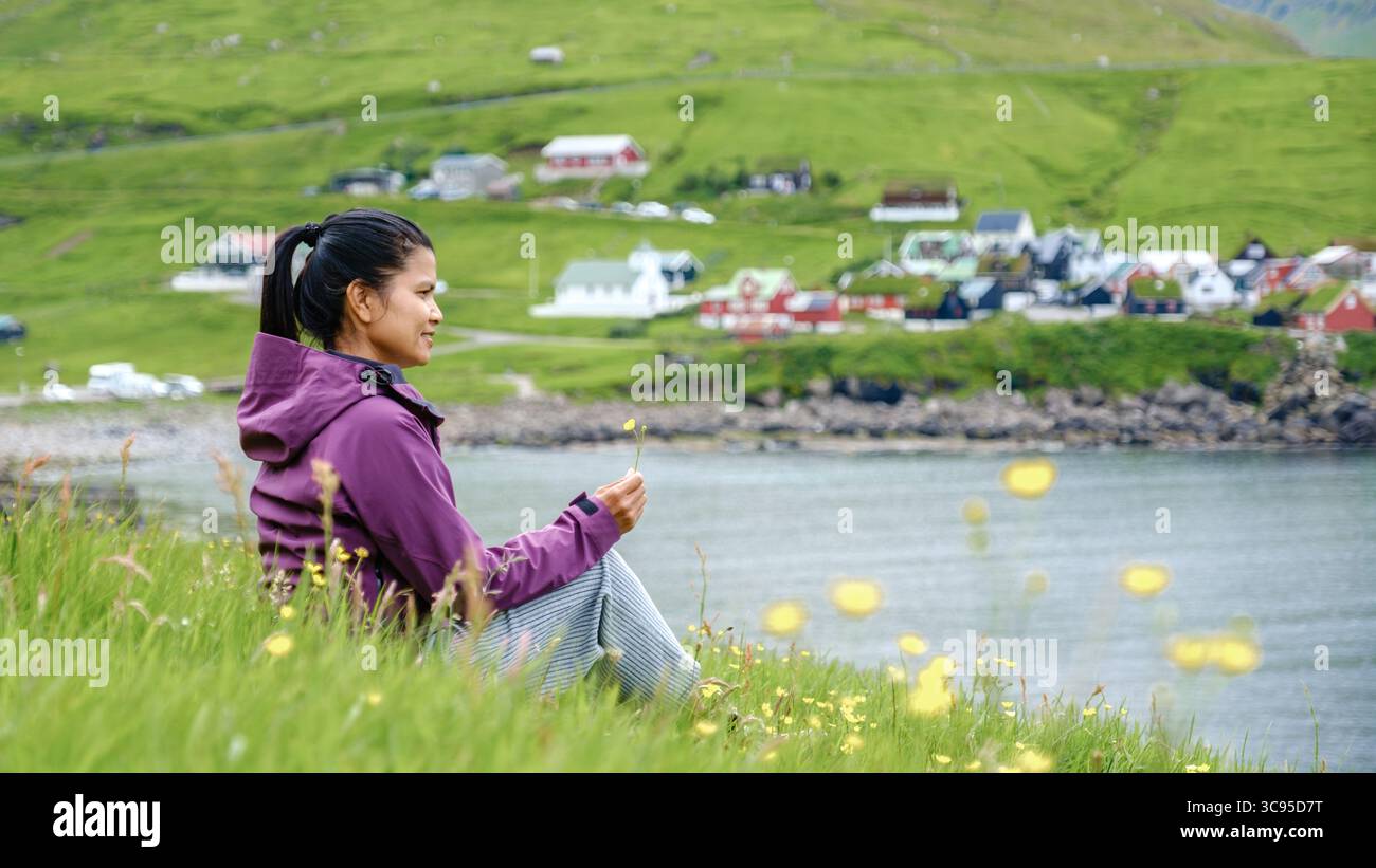 Eine Frau sitzt friedlich auf einem grasbewachsenen Hügel auf den Färöern, umgeben von lebendigen Wildblumen. Elduvik Village Stockfoto