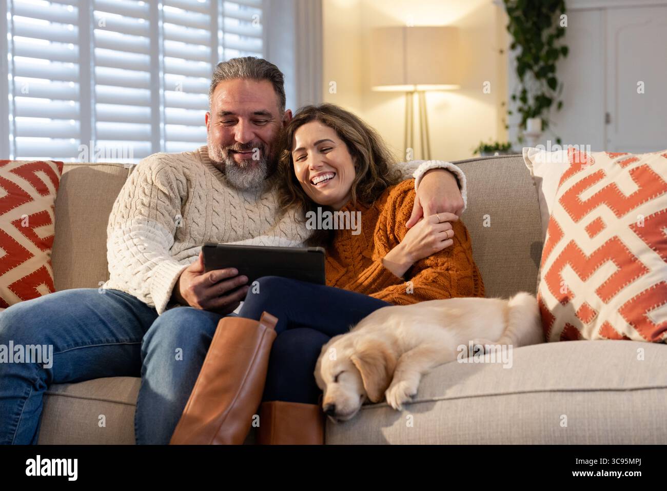 Reifes erwachsenes Paar, das sich auf einer beigen Couch in einem gemütlichen Wohnzimmer mit Hündchen lehnt und die Tablette hält Stockfoto