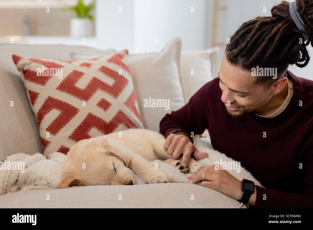 Afroamerikaner, der sich im Wohnzimmer auf der Couch stützt und den schlafenden Welpen auf der Decke beobachtet, im Kopierraum Stockfoto
