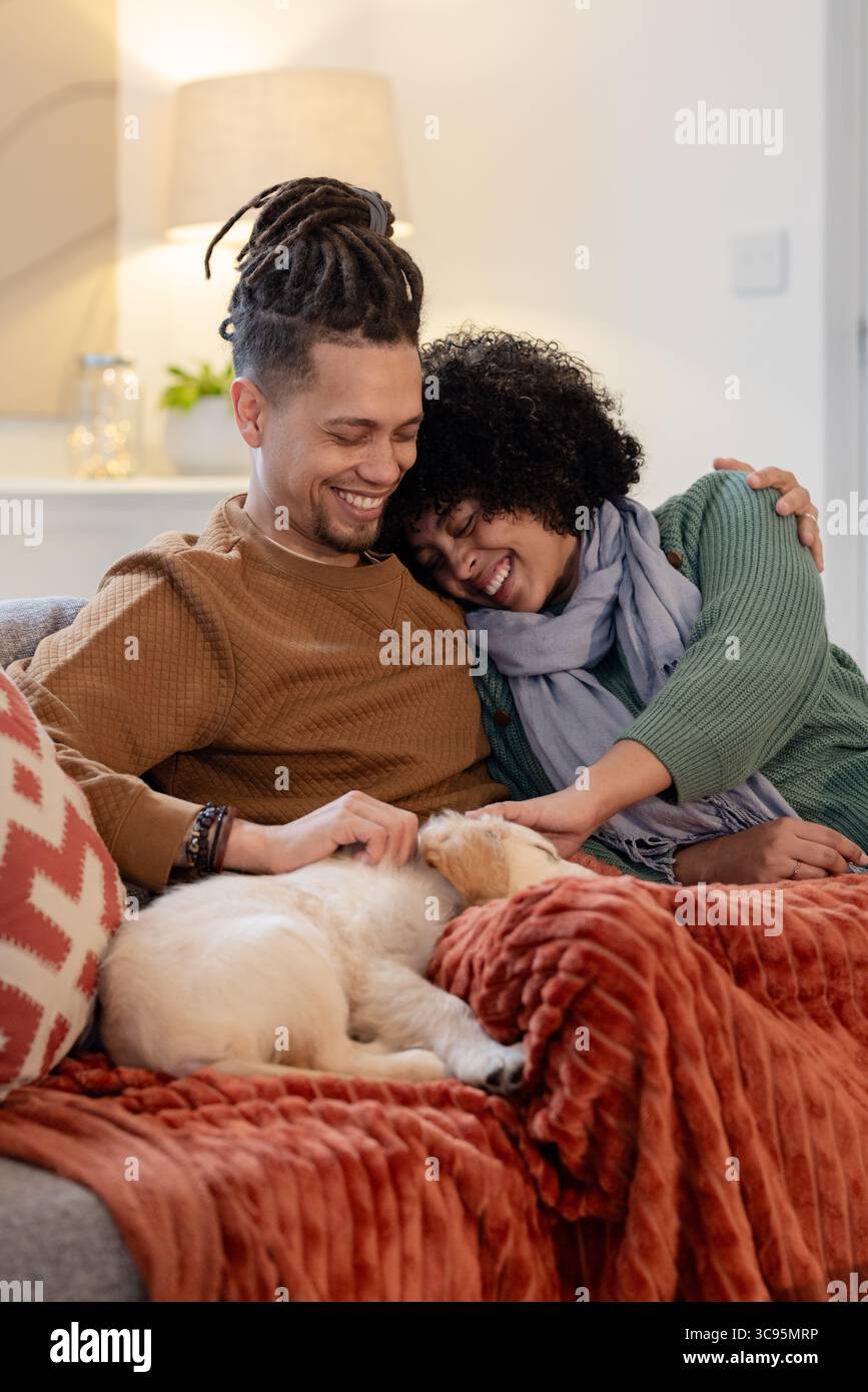 Afroamerikanisches Paar sitzt auf der Couch und streichelt weißen und braunen Welpen auf der Decke im Wohnzimmer Stockfoto