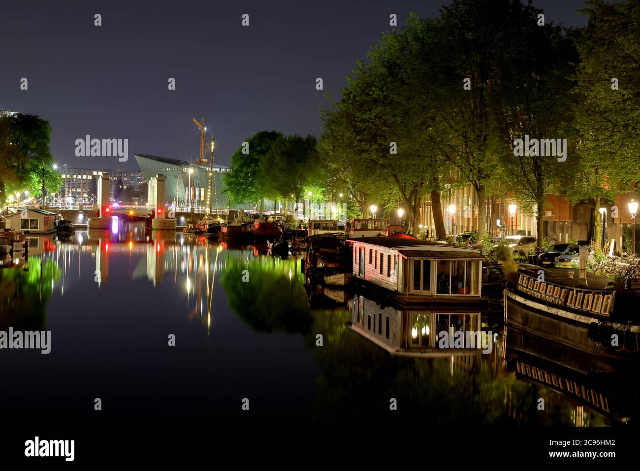 Die Lichter der Montelbaanstoren spiegeln sich im Oudeschans-Kanal wieder, wo historische Fassaden, vertäute Schiffe und Ampelpfosten am Fluss in der Amsterdamer Nachtszene leuchten. Stockfoto