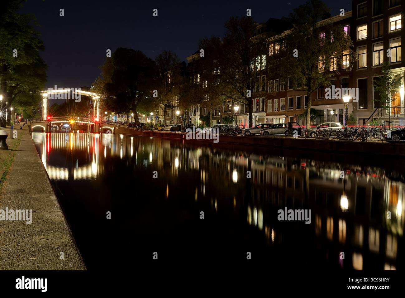 Nächtliches Panorama des Amsterdamer Walter Suskindbrug mit beleuchteter Brücke und Kanalreflektionen unter einem Halbmond, die einen ruhigen urbanen Charme erwecken. Stockfoto