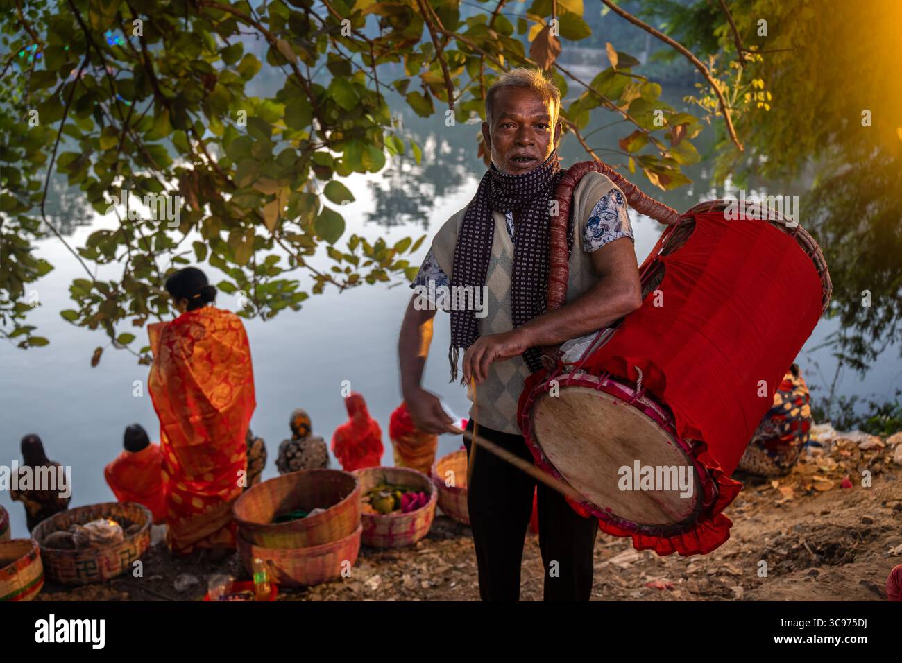 Bogura, Bangladesch - 20. November 2023: Blick auf einen Mann, der eine große, leuchtende rote Trommel in der Nähe eines Flusses spielt, wo sich Menschen mit Opfern versammeln. Stockfoto