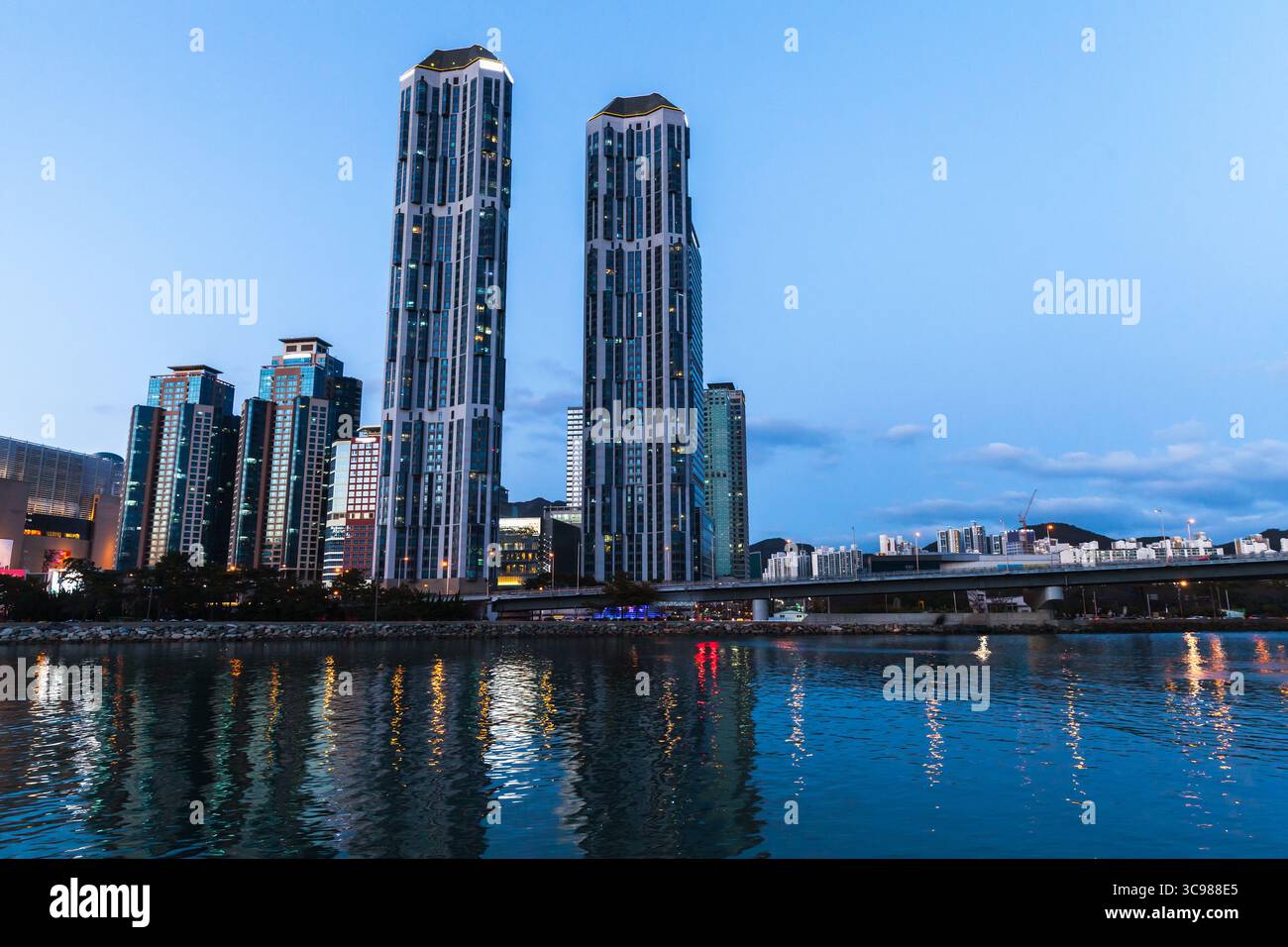 Die Skyline von Busan zeigt hohe, moderne Wolkenkratzer, die in der Abenddämmerung beleuchtet werden und sich auf dem ruhigen Wasser des Ufers spiegeln und von einem ruhigen Abendhimmel umgeben sind. Sout Stockfoto