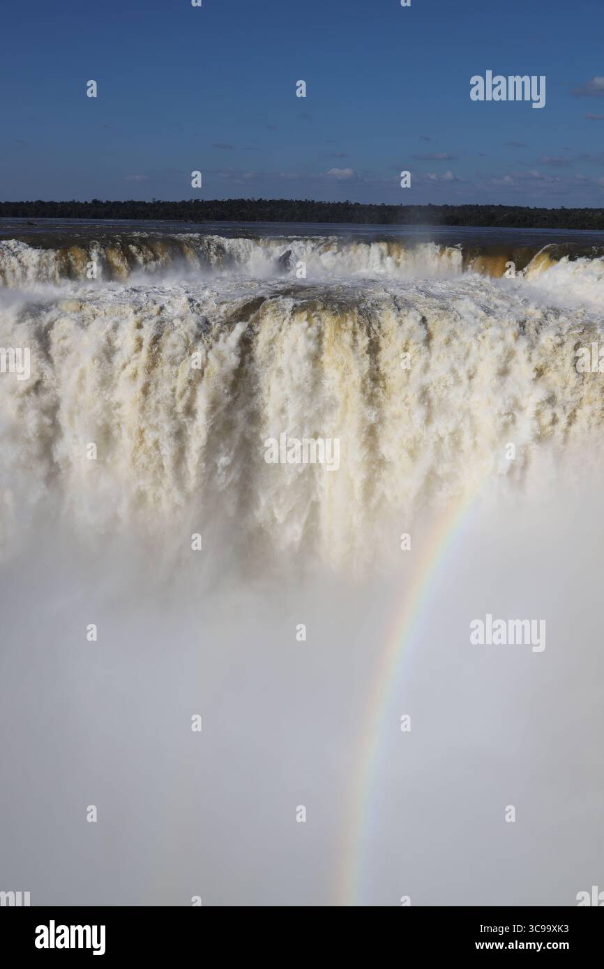 Atemberaubender Blick auf die Iguazú-Wasserfälle an der Teufelskehle (Garganta del Diablo) mit Regenbogen auf der argentinischen Seite, unter einem hellblauen Himmel an einem sonnigen Tag. Stockfoto