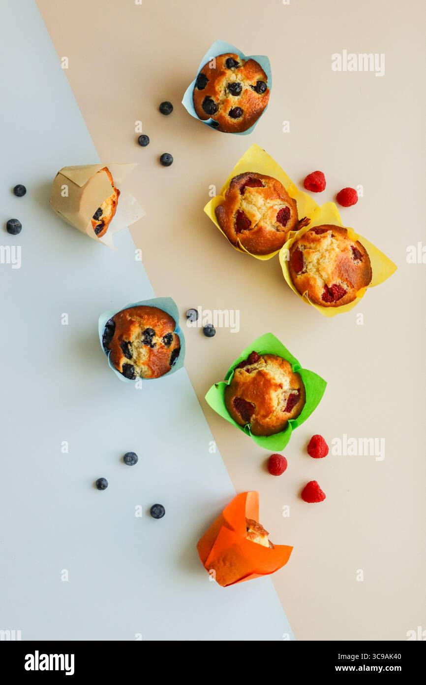 Verschiedene Beerenmuffins – Blaubeere und Himbeere – in farbenfrohen Tulpenpapiereinlagen in Blau-, Grün-, Gelb-, Orange- und neutralen Tönen gewickelt. Stockfoto