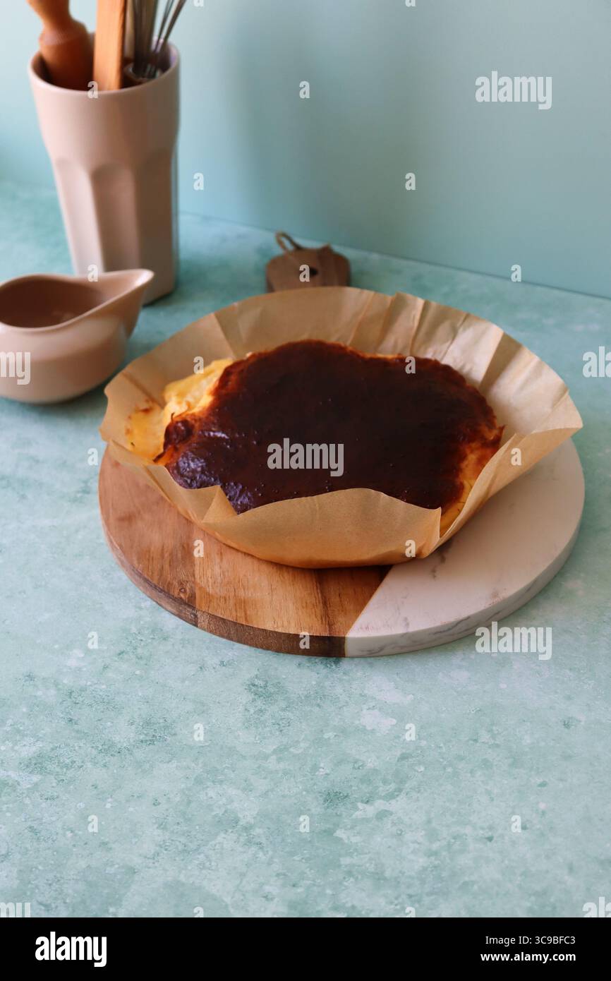 Ein frisch gebackener baskischer Käsekuchen, der in seinem zerknitterten Pergamentpapier auf einem runden Schneidebrett aus Holz und Marmor vor einer rustikalen Küche serviert wird Stockfoto