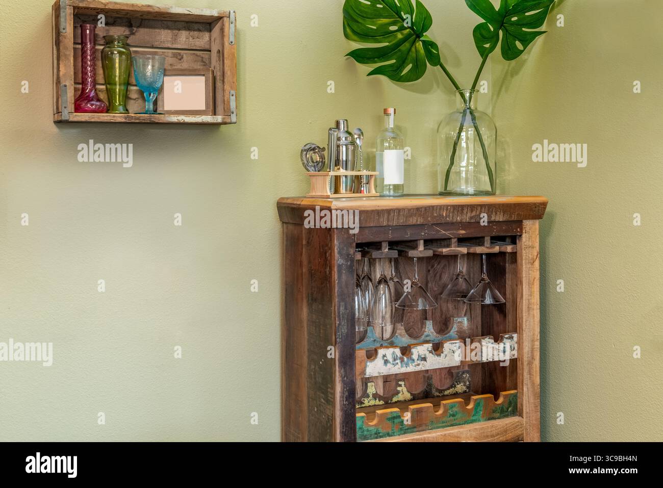 Rustikaler Barschrank aus Holz mit Trinkgläsern und einem kleinen Regal an einer hellgrünen Wand Stockfoto