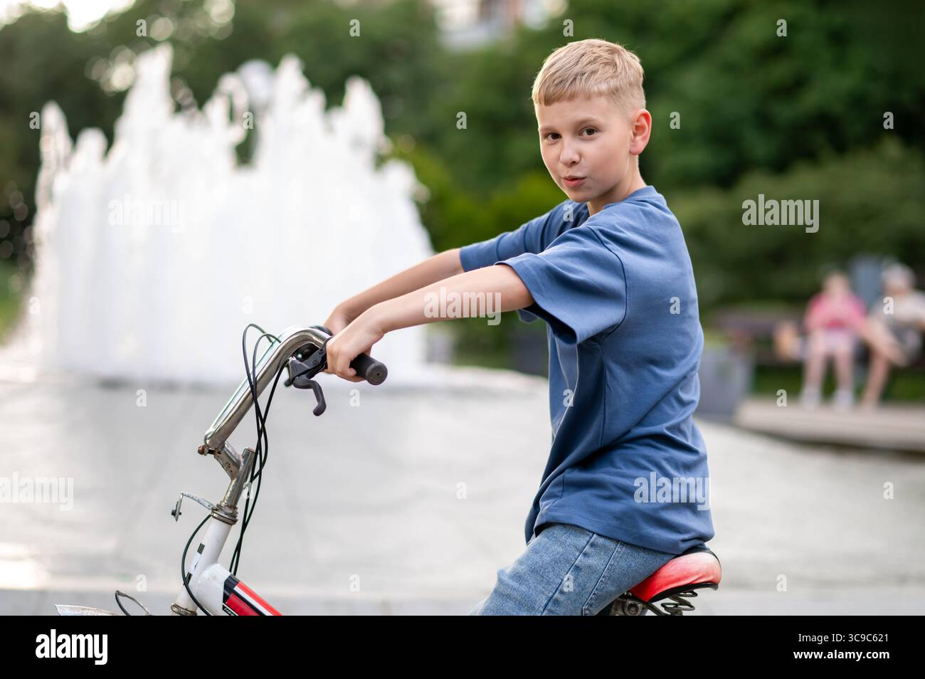 Der blonde Teen-Junge radelt an einem dekorativen Brunnen in einem Stadtpark an einem sonnigen Tag und genießt einen aktiven Outdoor-Lifestyle, ein Konzept von Jugend, Freiheit und Erholung Stockfoto