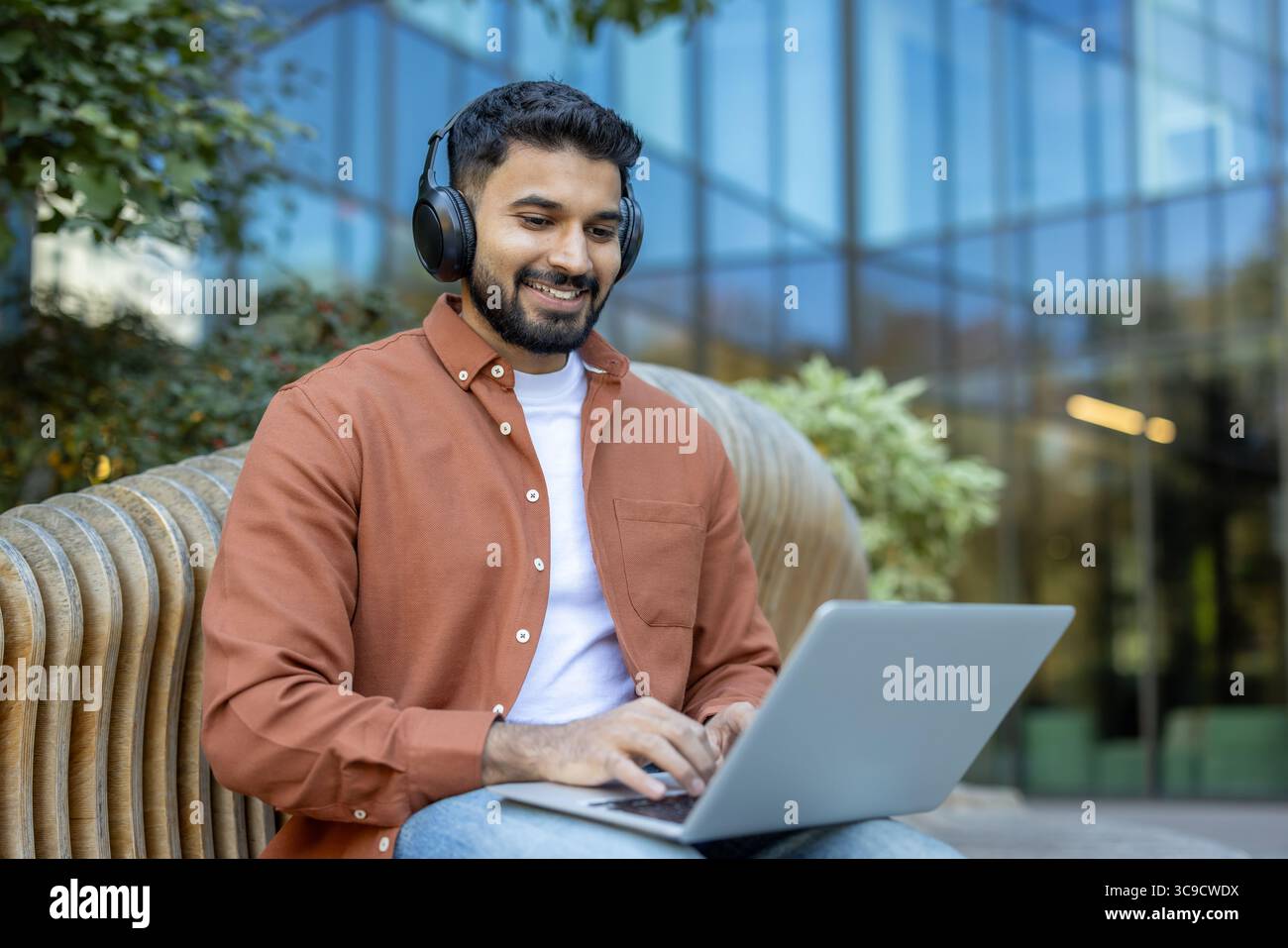 Ein Indianer, der Kopfhörer trägt, lächelt, während er draußen auf einer Bank einen Laptop benutzt, umgeben von Grün. Stockfoto