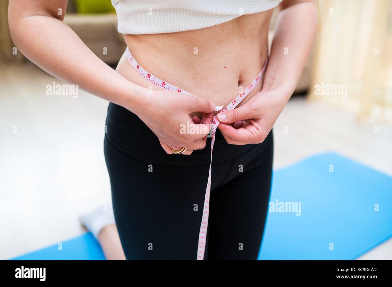 Frau mit unvollkommener Taille, die sie mit Klebeband misst, während sie auf Yogamatte steht, Körperbewusstsein und Selbstpflegekonzept Stockfoto