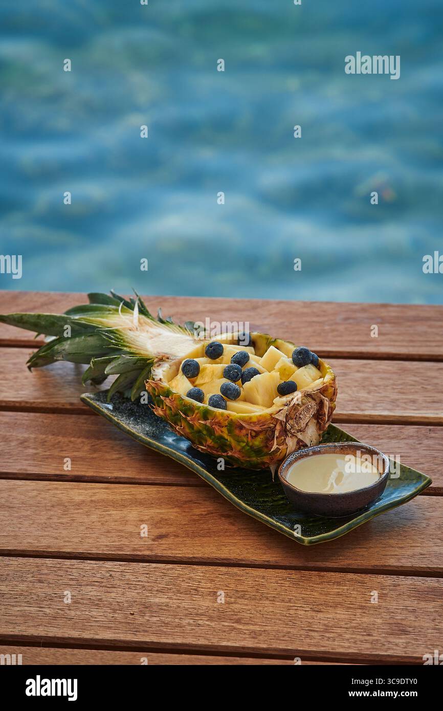 Frisch geschnittene Ananas gefüllt mit saftigen gelben Früchten und Heidelbeeren, serviert auf einem natürlichen Holztisch am sprudelnden Wasser des Ozeans Stockfoto