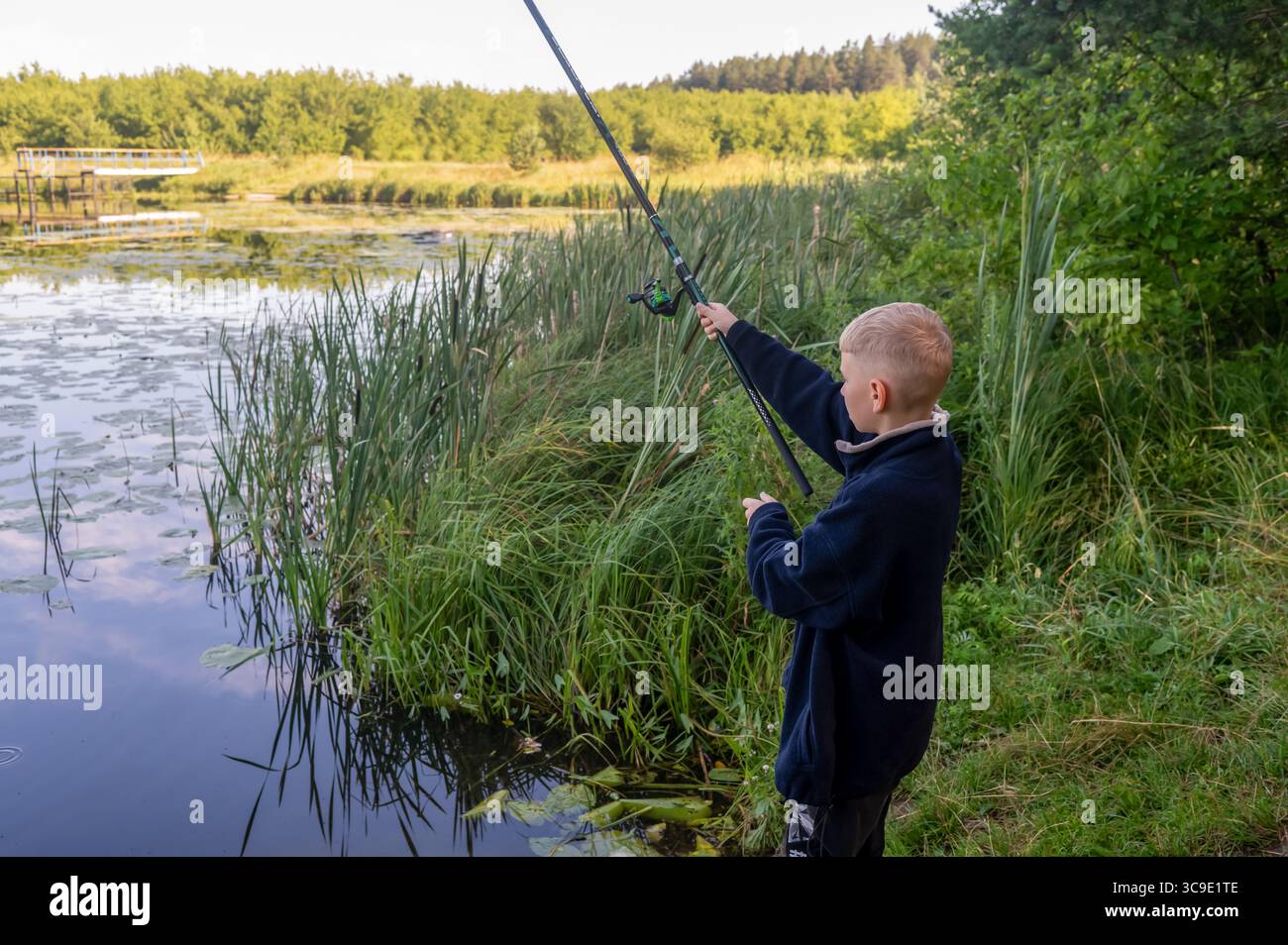 Fröhliches Kind, das am sonnigen Sommertag mit einer erhöhten Angelrute in der Nähe des Flusses erfolgreich fischt Stockfoto