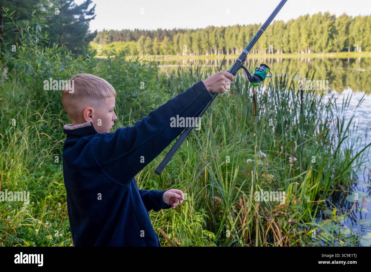 Fröhliches Kind, das am sonnigen Sommertag mit einer erhöhten Angelrute in der Nähe des Flusses erfolgreich fischt Stockfoto
