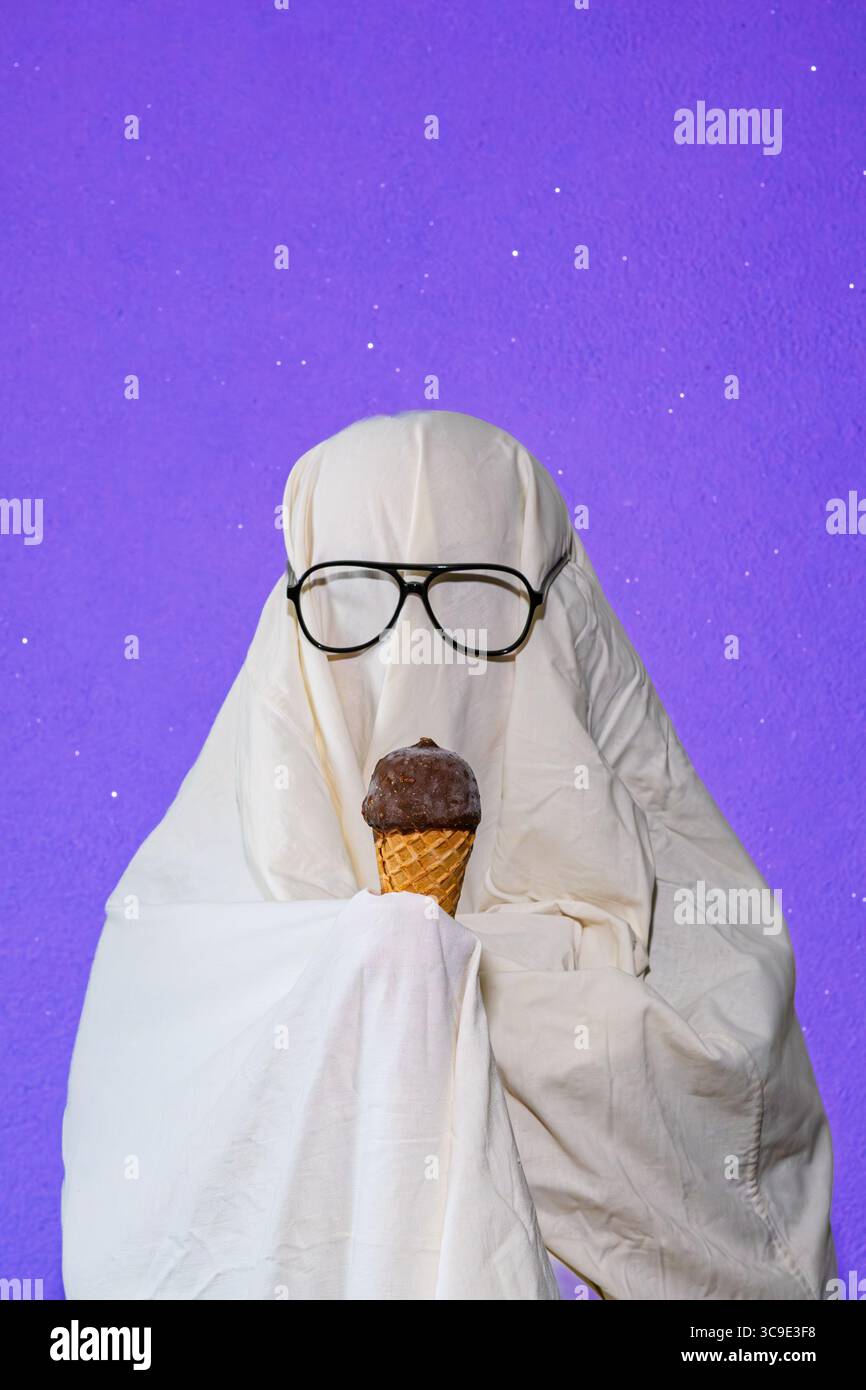 Ein humorvolles Bild einer Person, die in einem einfachen Geisterkostüm aus weißem Bettlaken gekleidet ist, Sonnenbrille trägt und eine Eiscreme isst. Erfasst Stockfoto