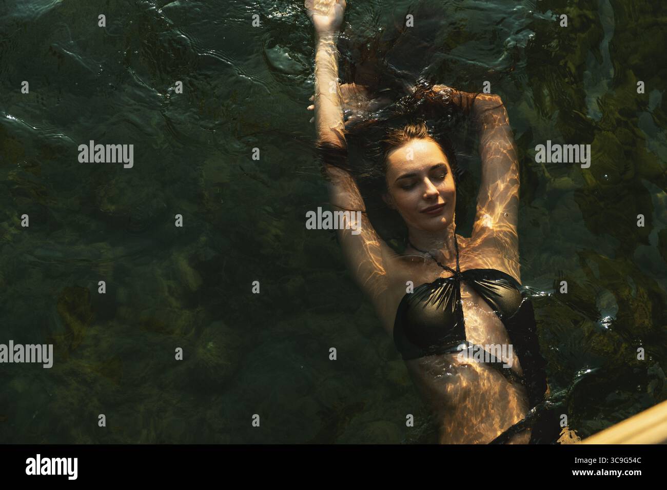 Atemberaubende Vogelperspektive einer brünetten Frau, die sich im Meer entspannt, in schwarzem Bikini unter goldenem Sonnenuntergang. Stockfoto