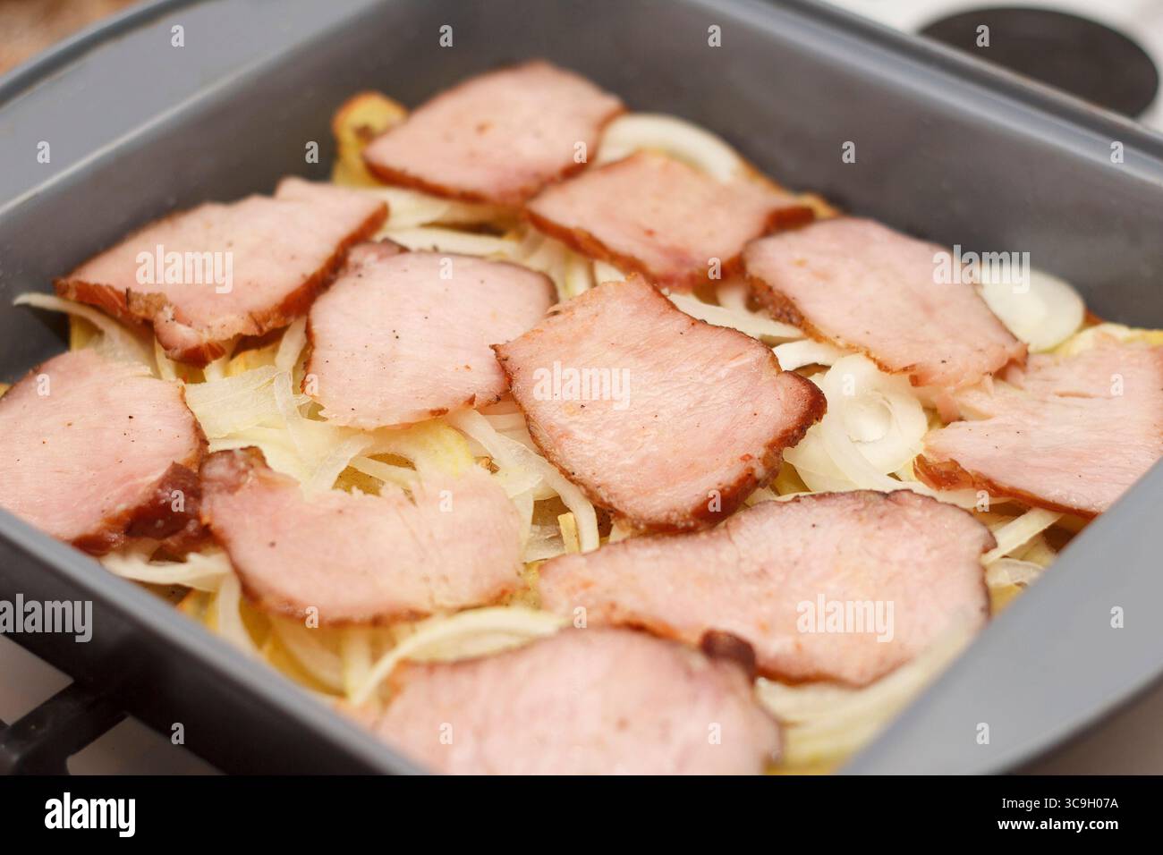In Scheiben geschnittener Schinken auf einem Bett mit Kartoffeln und Zwiebeln in einer Auflaufform. Das Gericht ist fertig zum Kochen und bietet eine herzhafte Mahlzeit Stockfoto