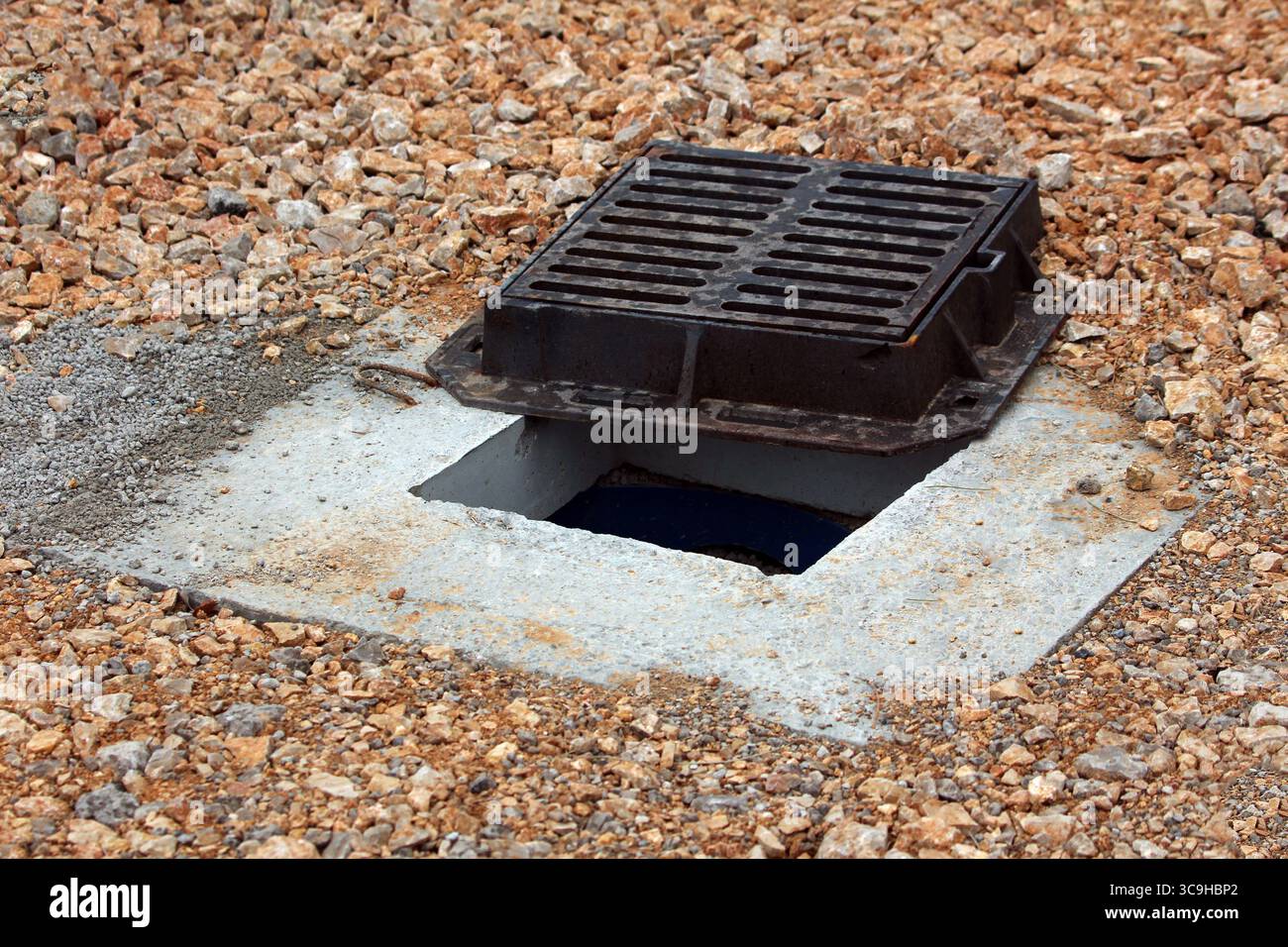 Ein schwarzes Metallrost ist leicht auf einem kiesbedeckten Boden offen und zeigt eine dunkle Entwässerungsöffnung unter einem Betonrahmen Stockfoto