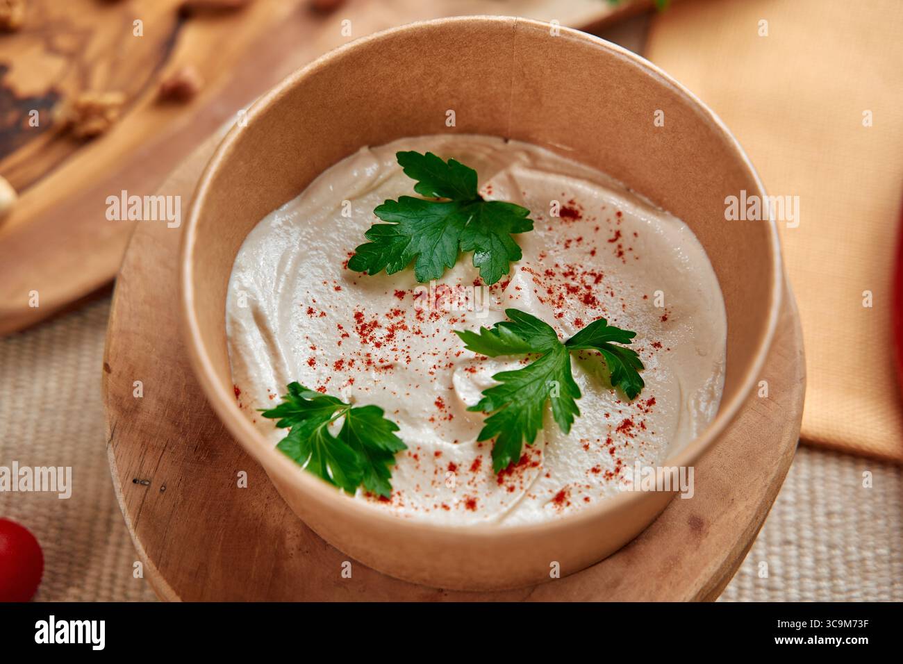 Köstlicher cremiger Dip garniert mit frischer Petersilie und Paprika, serviert in einer rustikalen Schüssel auf einem Holzbrett umgeben von frischen Zutaten Stockfoto