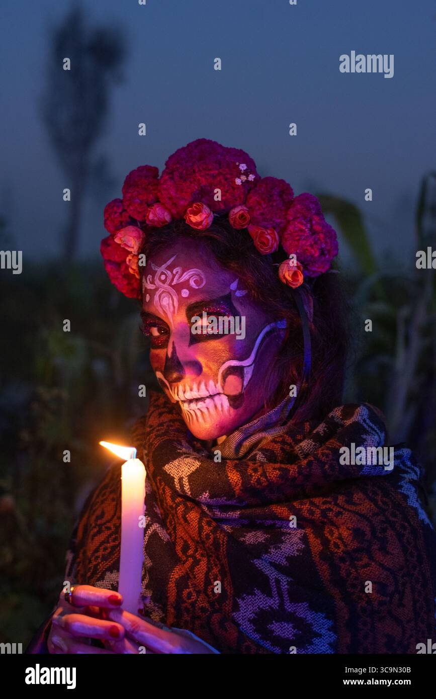 Eine Frau mit traditionellem Catrina-Make-up und einer Blumenkrone hält am frühen Morgen des Día de Muertos in Xochimilco, Mexiko, eine Kerze. Stockfoto
