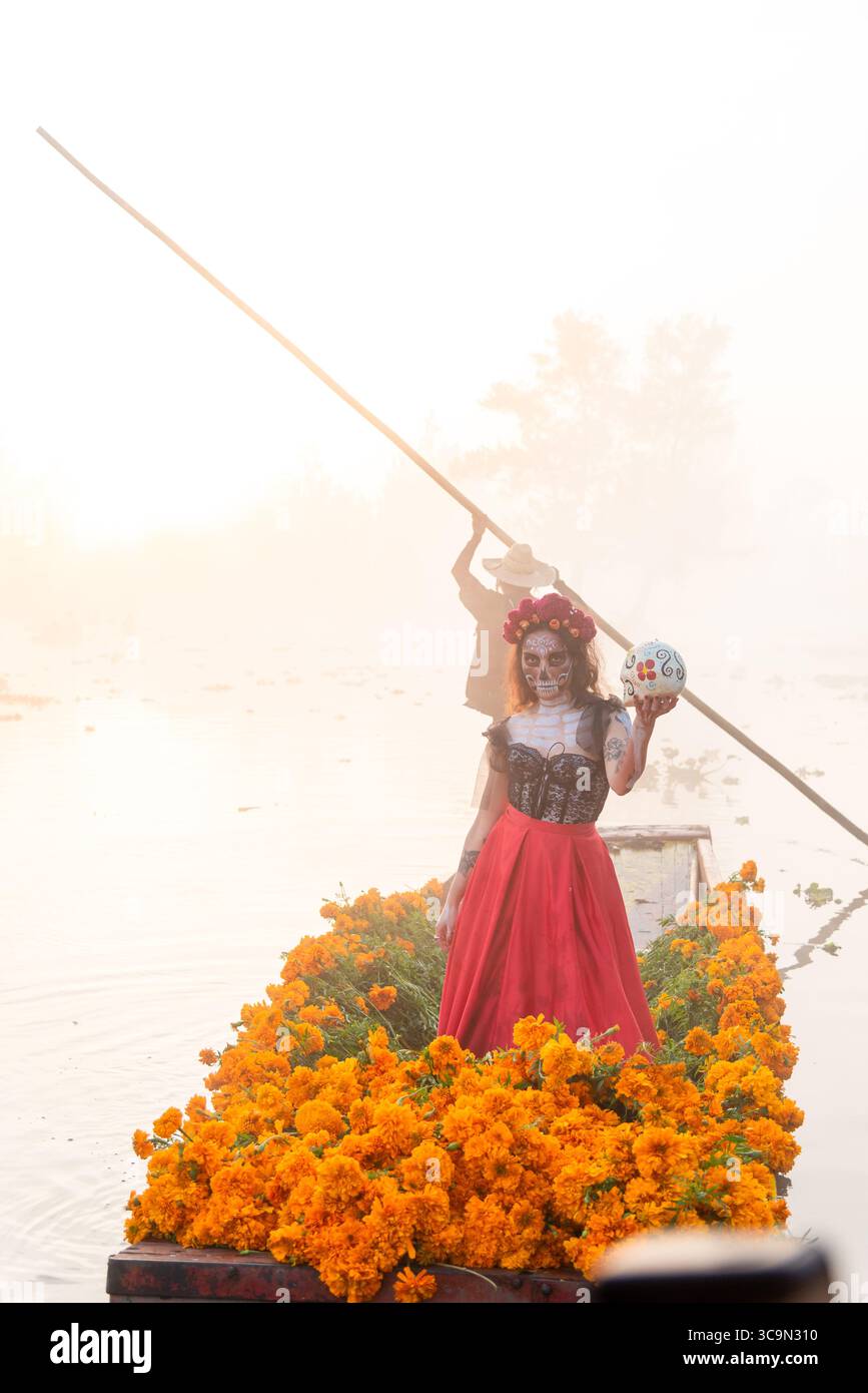 Frau, gekennzeichnet als La Catrina auf einem Trajinera-Boot mit Cempasuchil-Blüten während des Sonnenaufgangs in den Kanälen von Xochimilco, Mexiko. Stockfoto