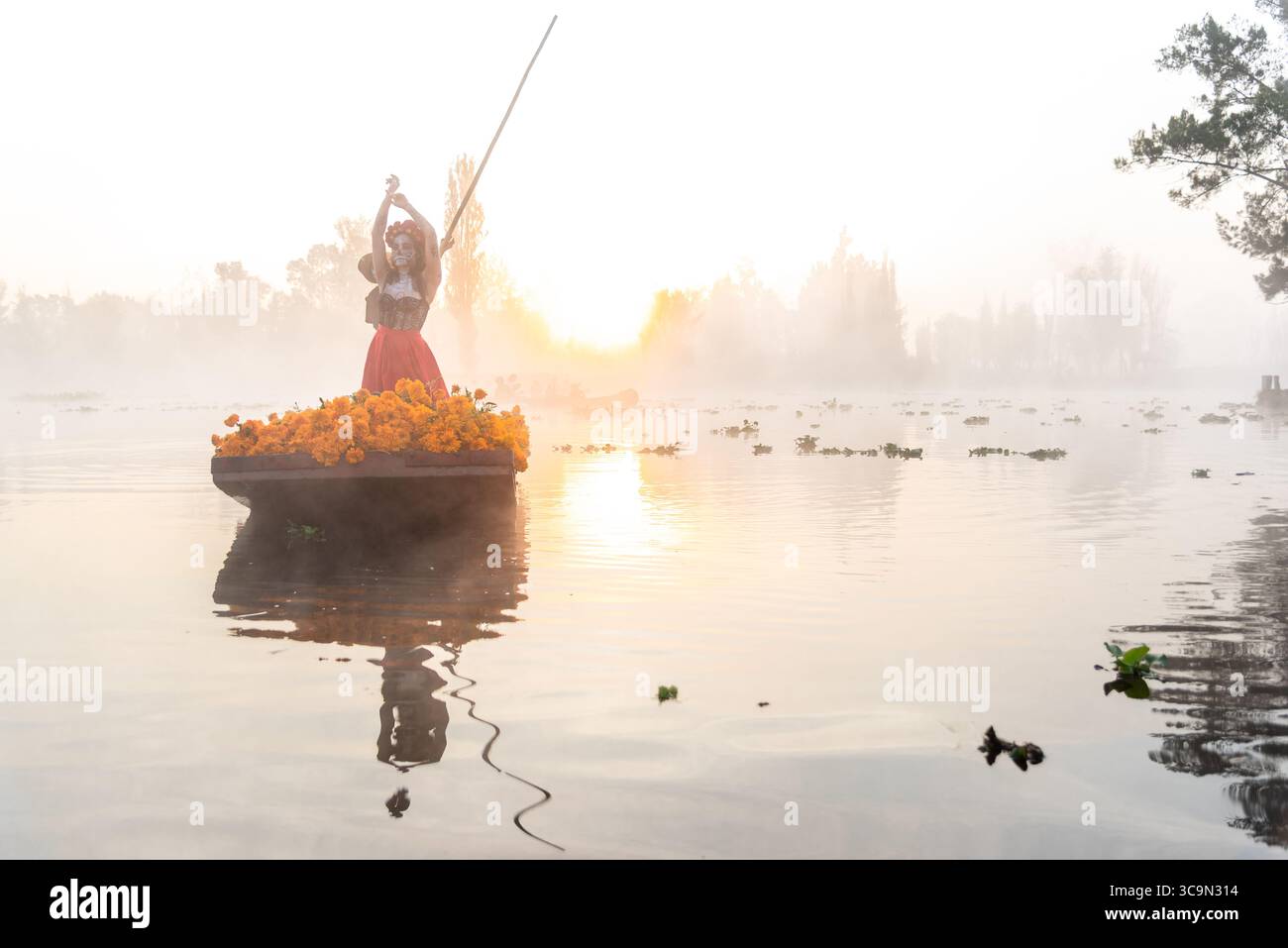 Frau, gekennzeichnet als La Catrina auf einem Trajinera-Boot mit Cempasuchil-Blüten während des Sonnenaufgangs in den Kanälen von Xochimilco, Mexiko. Stockfoto