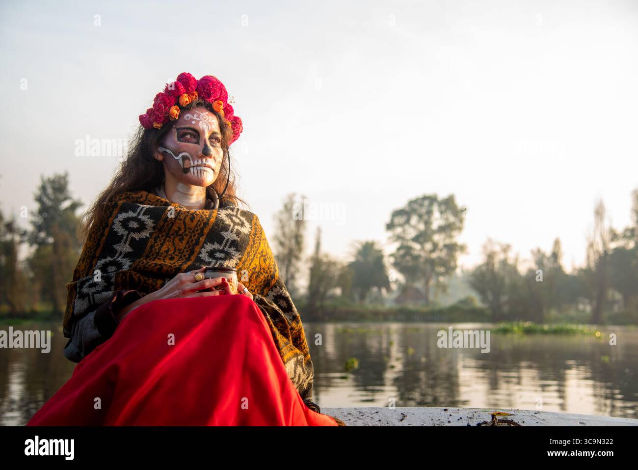 Eine Frau, die als La Catrina charakterisiert ist, auf einem Trajinera-Boot während des Sonnenaufgangs in den Kanälen von Xochimilco, Mexiko. Stockfoto