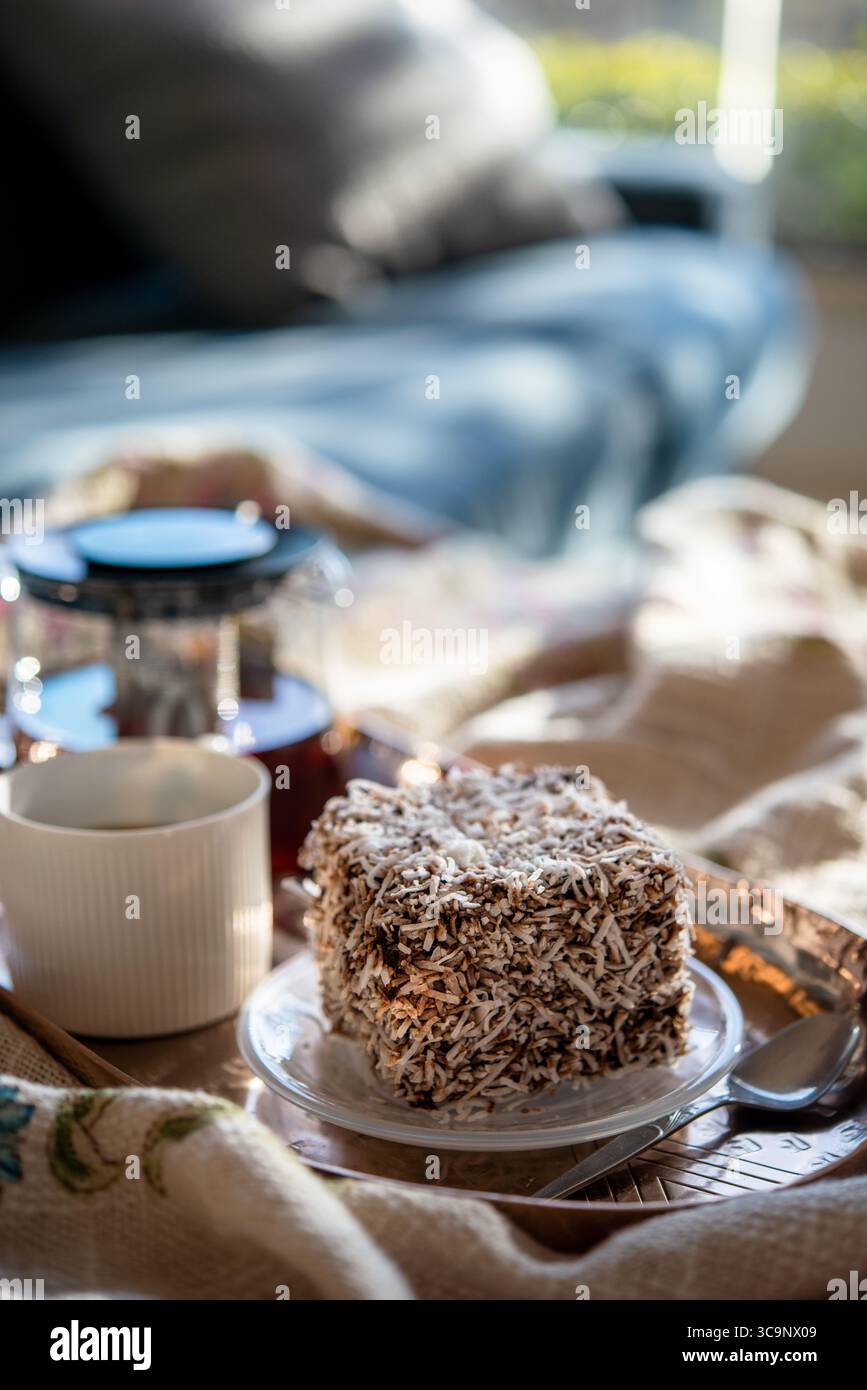 Ein köstlicher lamington-Kuchen ist perfekt auf einem Kupfertablett eingerahmt, mit einer Tasse Tee und einer Teekanne im Hintergrund. Stockfoto