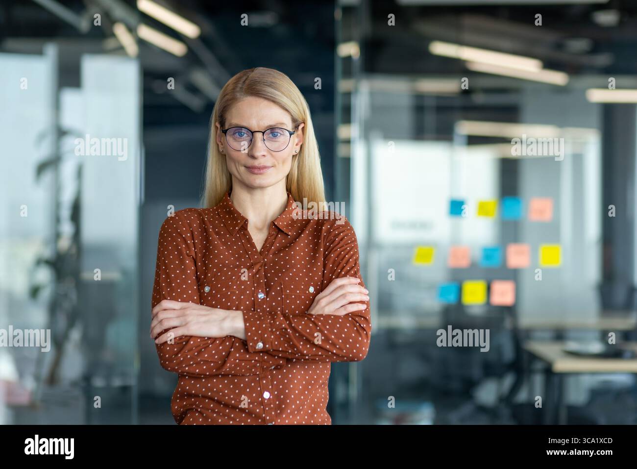 Eine blonde Frau in Brille mit überkreuzten Armen steht in einem Büro mit selbstbewusstem Ausdruck. Stockfoto