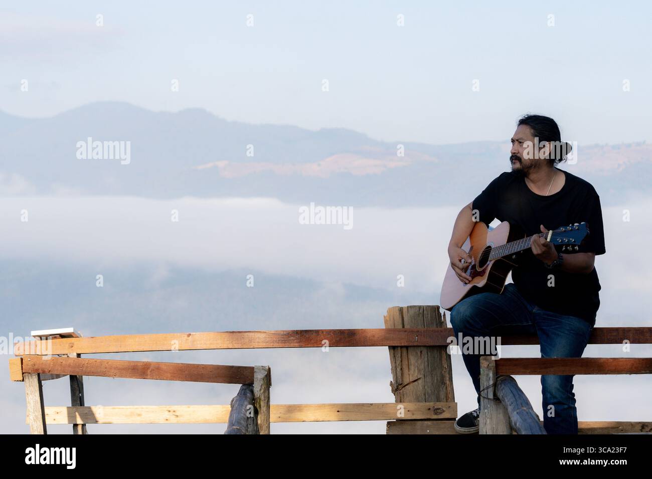 Junger Mann, der einen ruhigen Morgen genießt und Gitarre auf einem Holzbalkon mit warmem Sonnenlicht und malerischem Blick auf die Berge spielt. Stockfoto