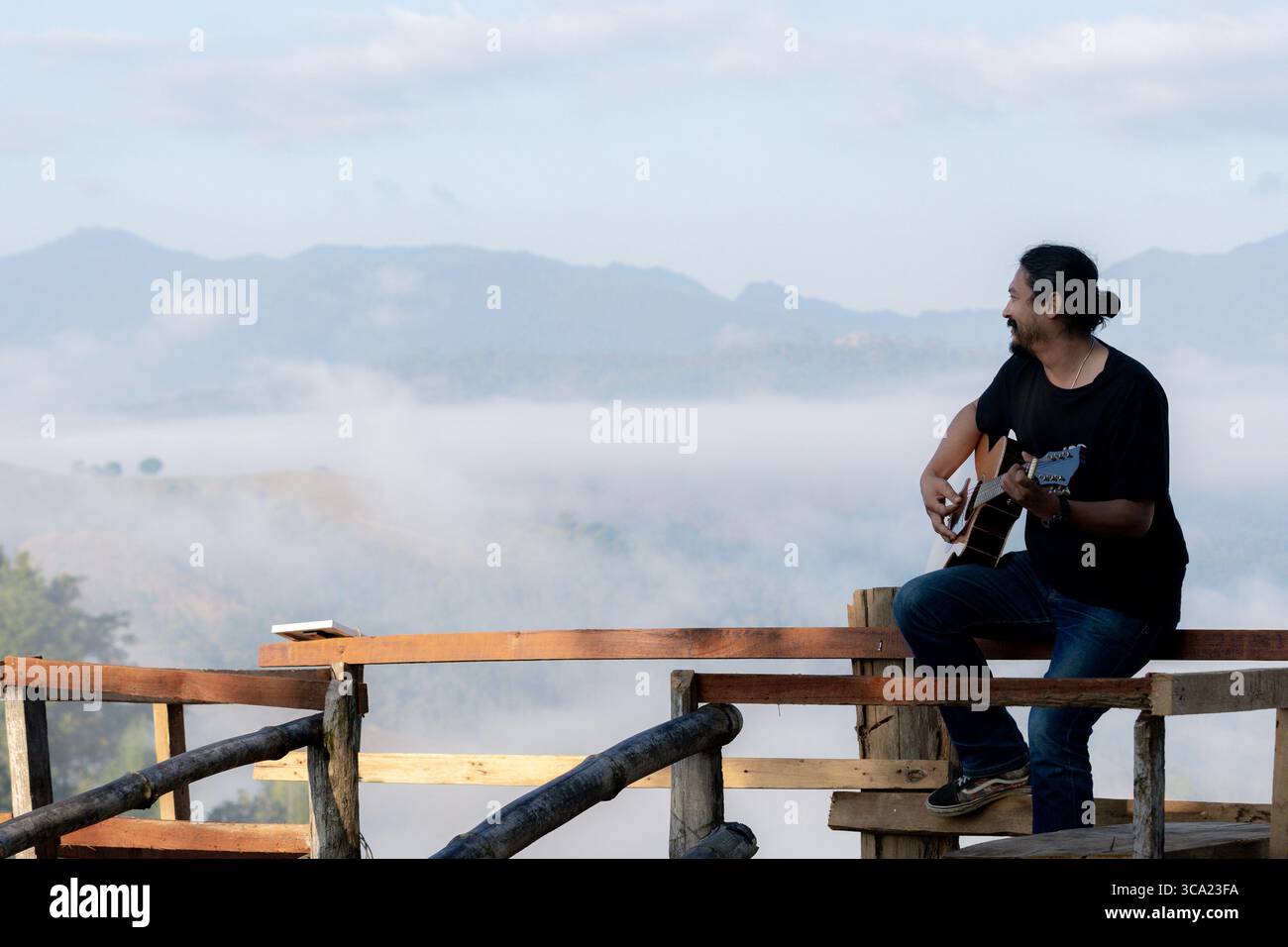 Junger Mann, der einen ruhigen Morgen genießt und Gitarre auf einem Holzbalkon mit warmem Sonnenlicht und malerischem Blick auf die Berge spielt. Stockfoto