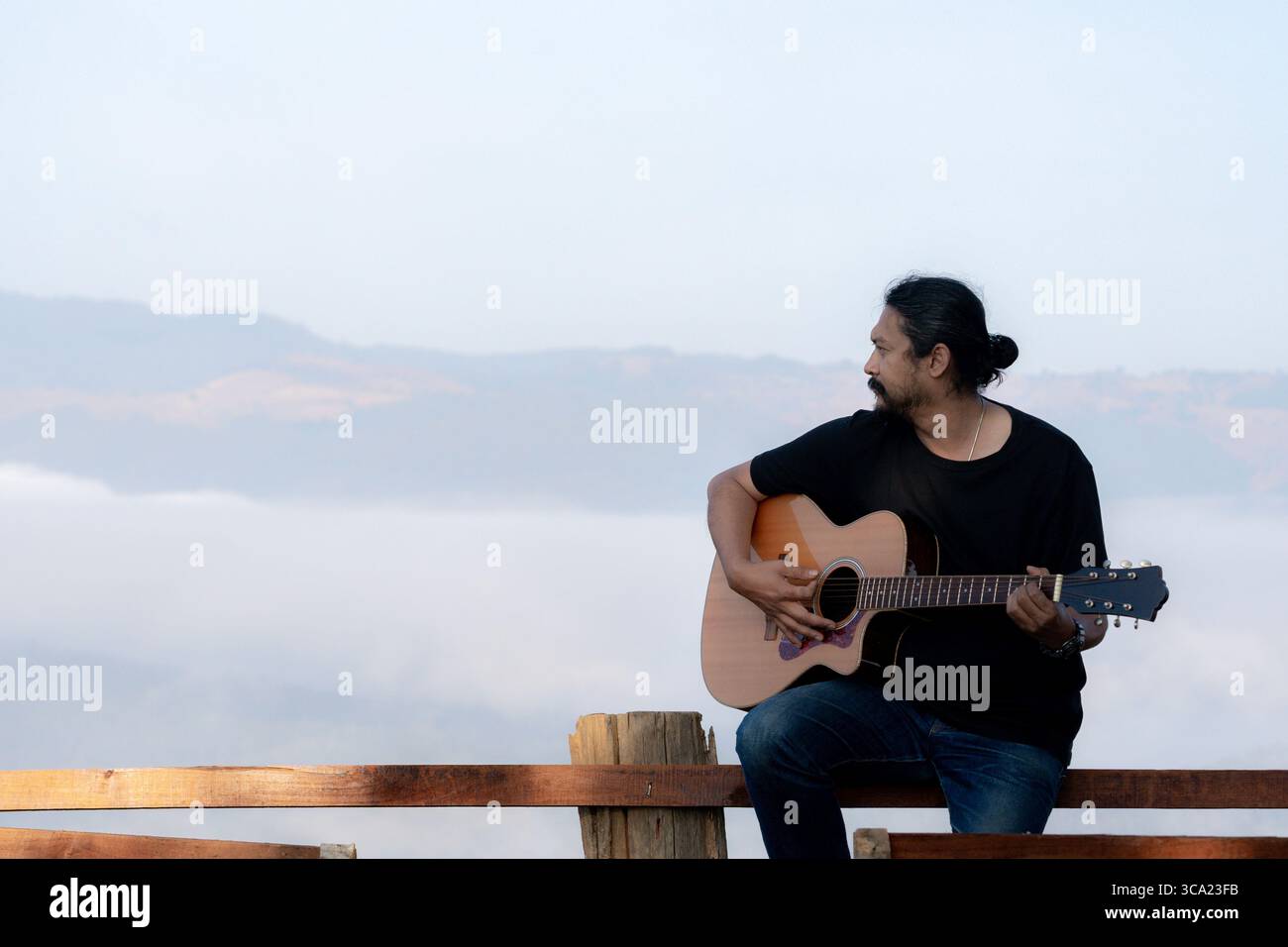 Junger Mann, der einen ruhigen Morgen genießt und Gitarre auf einem Holzbalkon mit warmem Sonnenlicht und malerischem Blick auf die Berge spielt. Stockfoto
