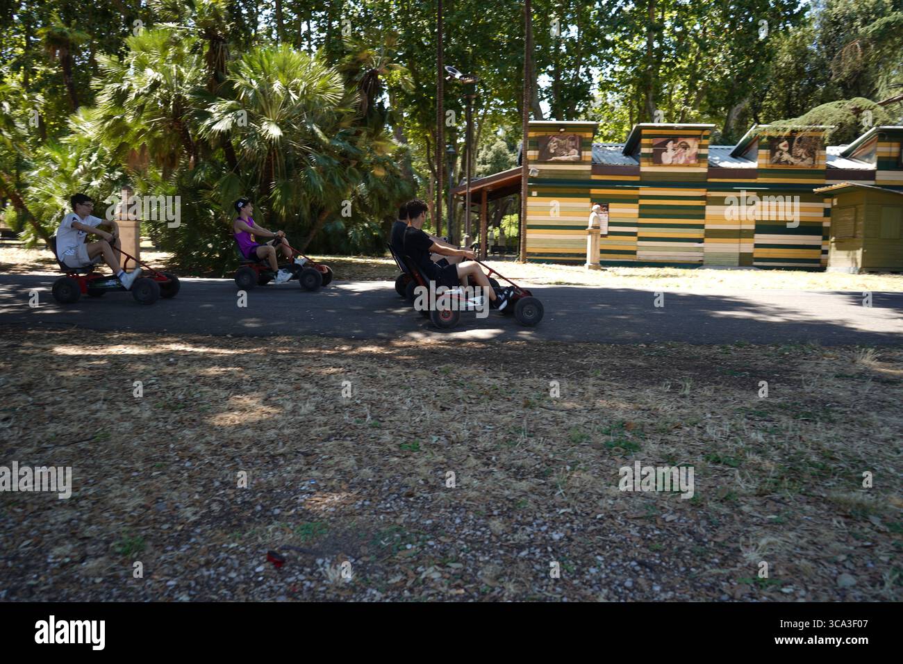 Drei Personen fahren mit Gokarts auf einem gepflasterten Weg vorbei an der Casa del Cinema, einem farbenfrohen Gebäude im Park der Villa Borghese, und genießen einen sonnigen Tag. Stockfoto