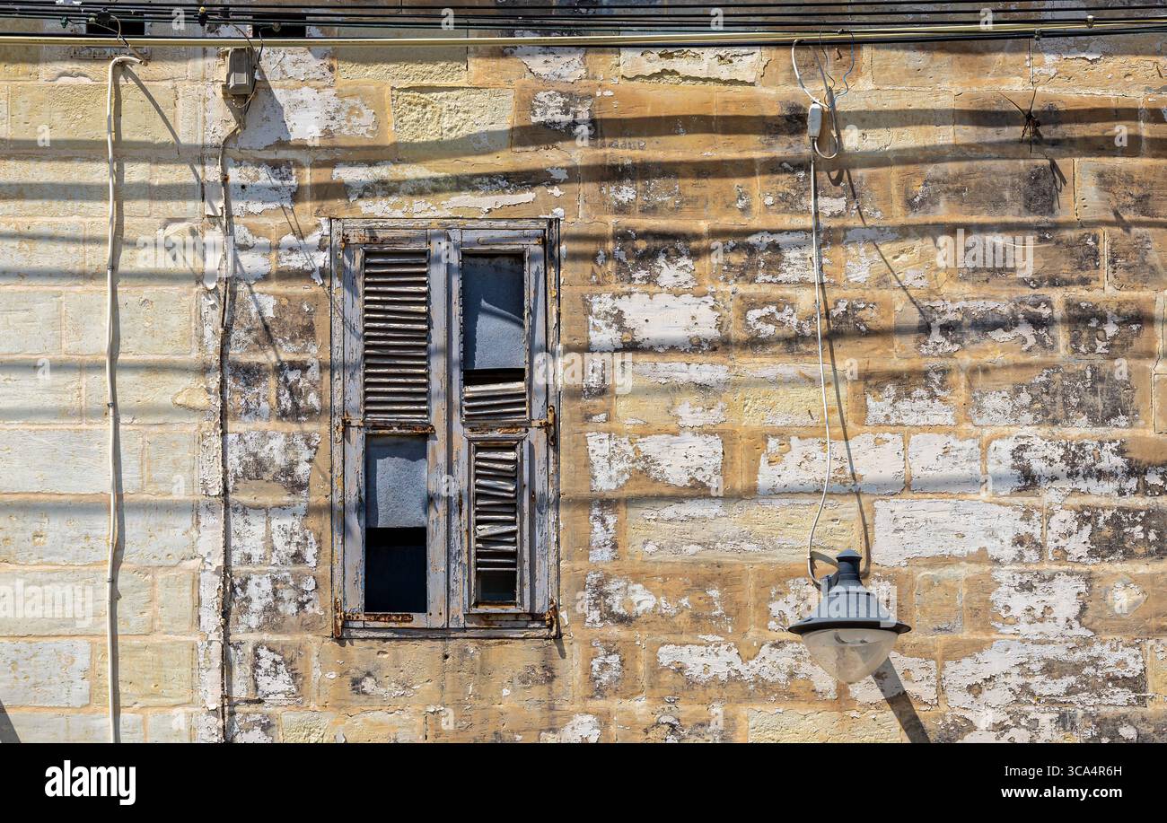 Schatten, die von Freileitungen auf das alte Kalksteingebäude geworfen werden. Zerbrochene und erodierte Holzfenster, Marsaxlokk, Malta Stockfoto