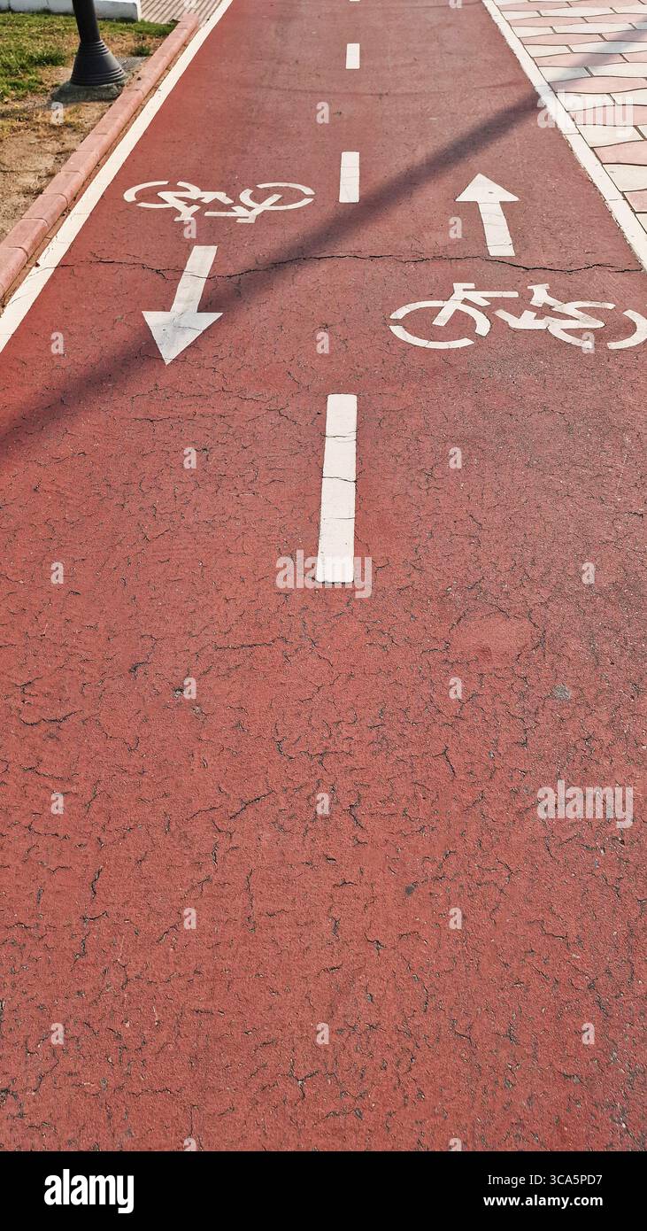 Almeria, Spanien - 25. Mai 2025: Radweg mit Richtungspfeilen und Fahrradsymbolen auf gerissenem rotem Gehweg, umgeben von grünem Gras und urbanem Gelände Stockfoto