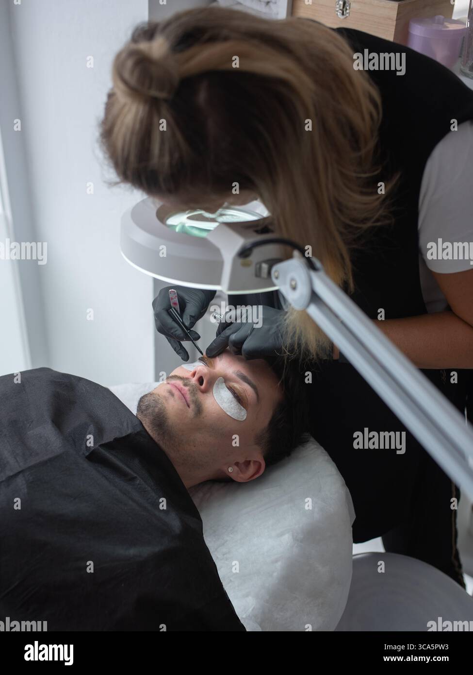 Die Kosmetikerin wendet geschickt Wimpernverlängerungen an männliche Kunden in einem modernen Salon an und betont die Liebe zum Detail und die professionelle Atmosphäre Stockfoto