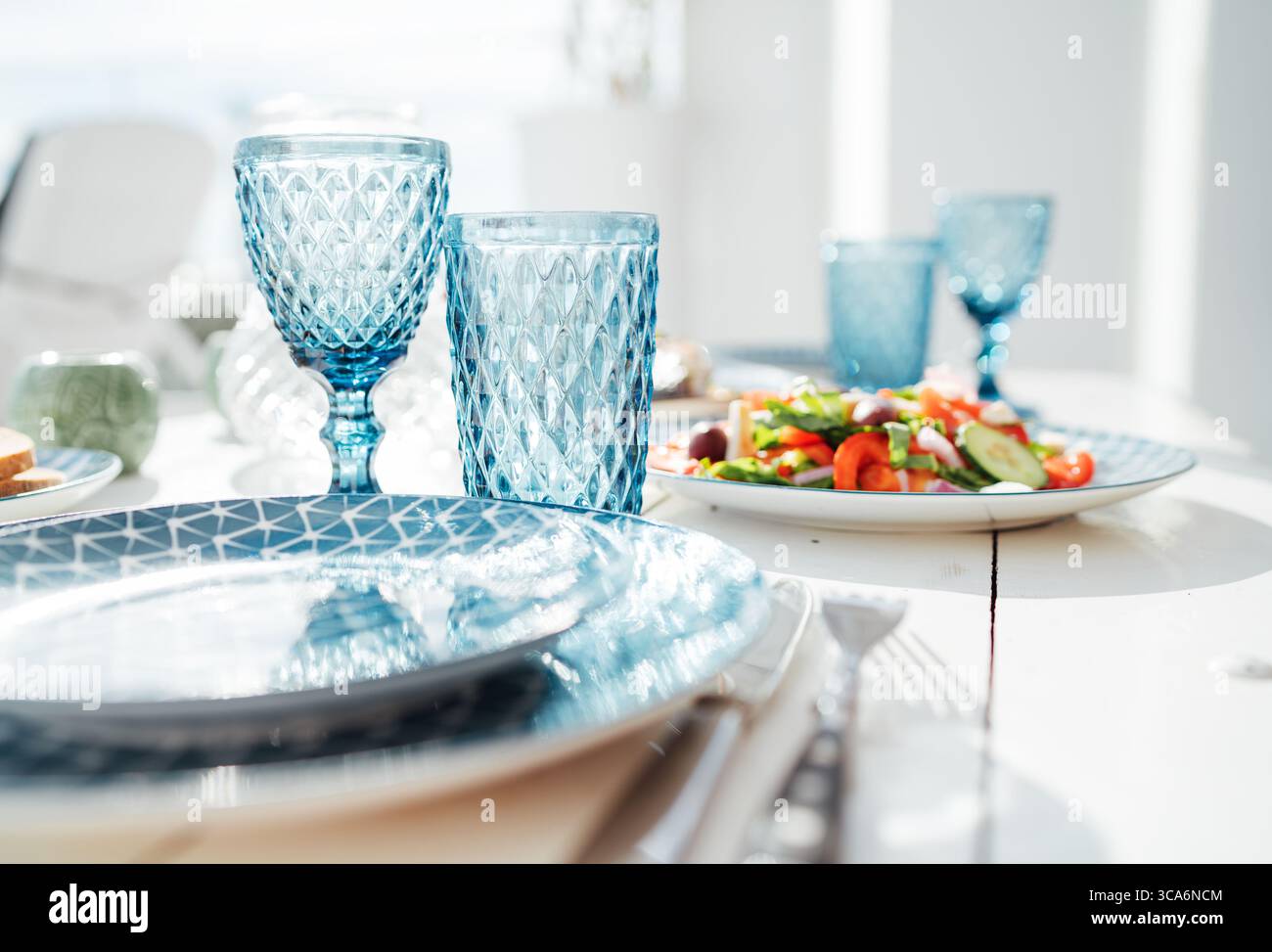 Eleganter Sommertisch mit gemusterten blauen Gläsern, Keramikplatten, frischem Gemüsesalat und weichem Tageslicht für eine gemütliche, einladende Atmosphäre Stockfoto